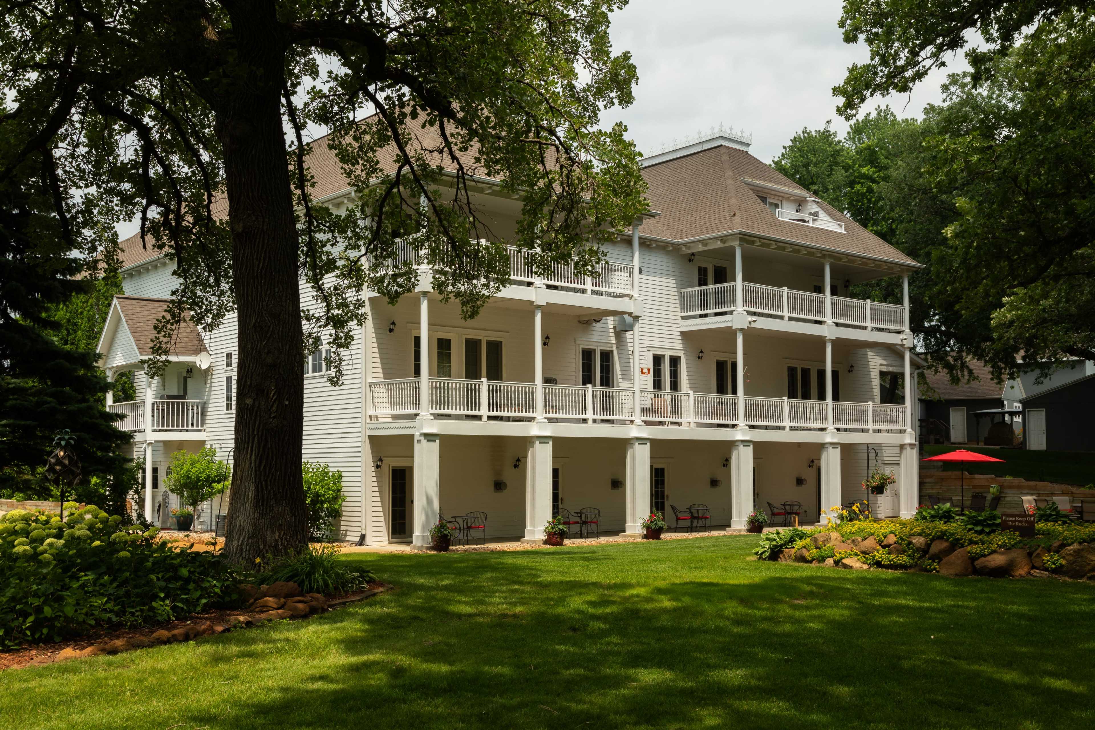 A large, two-story white house with multiple balconies stands in a grassy area surrounded by trees and landscaped gardens.