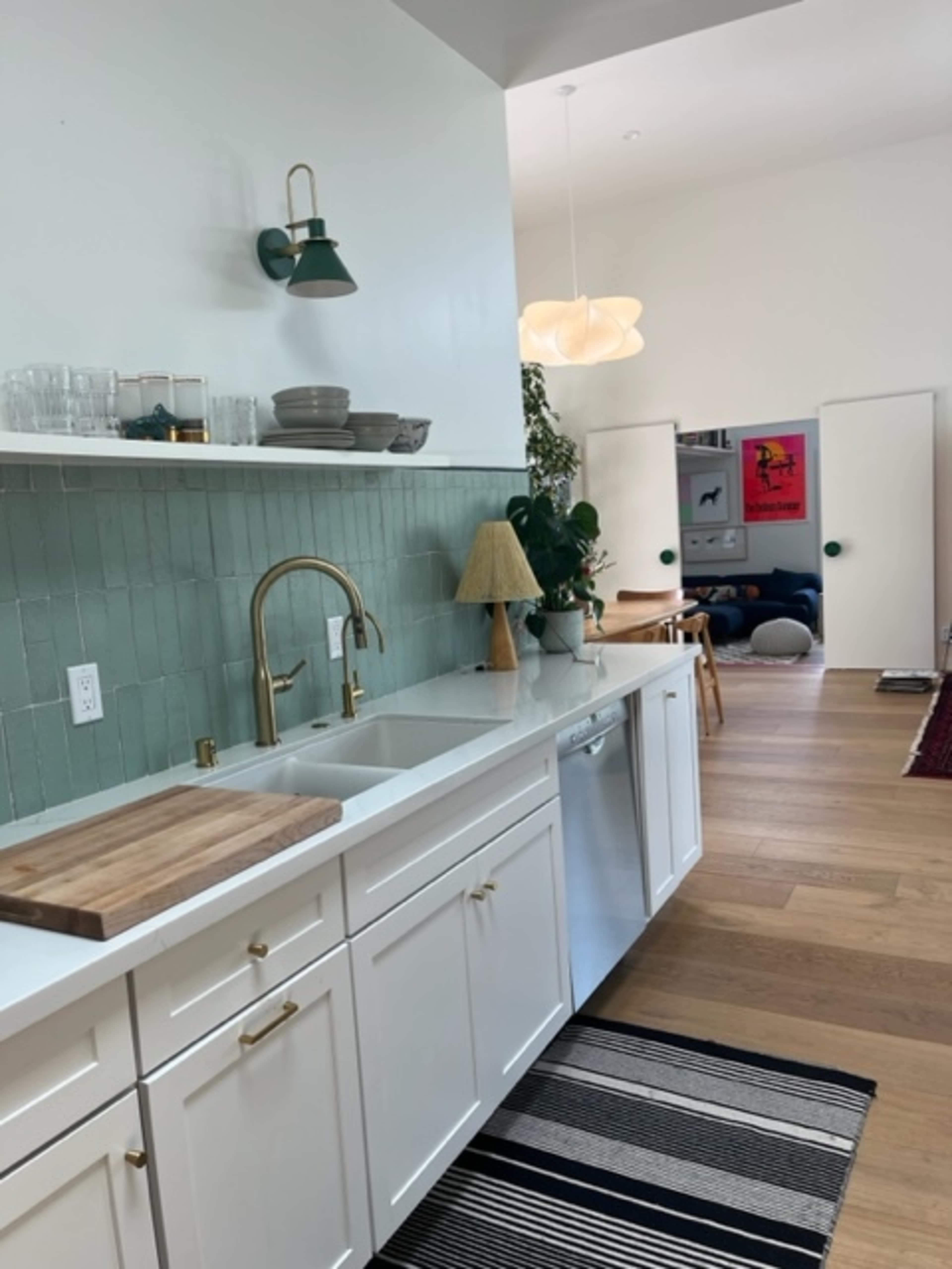 A modern kitchen with white cabinetry, a green tiled backsplash, a wooden cutting board on the counter, and a view into a living area featuring a colorful poster on the wall.