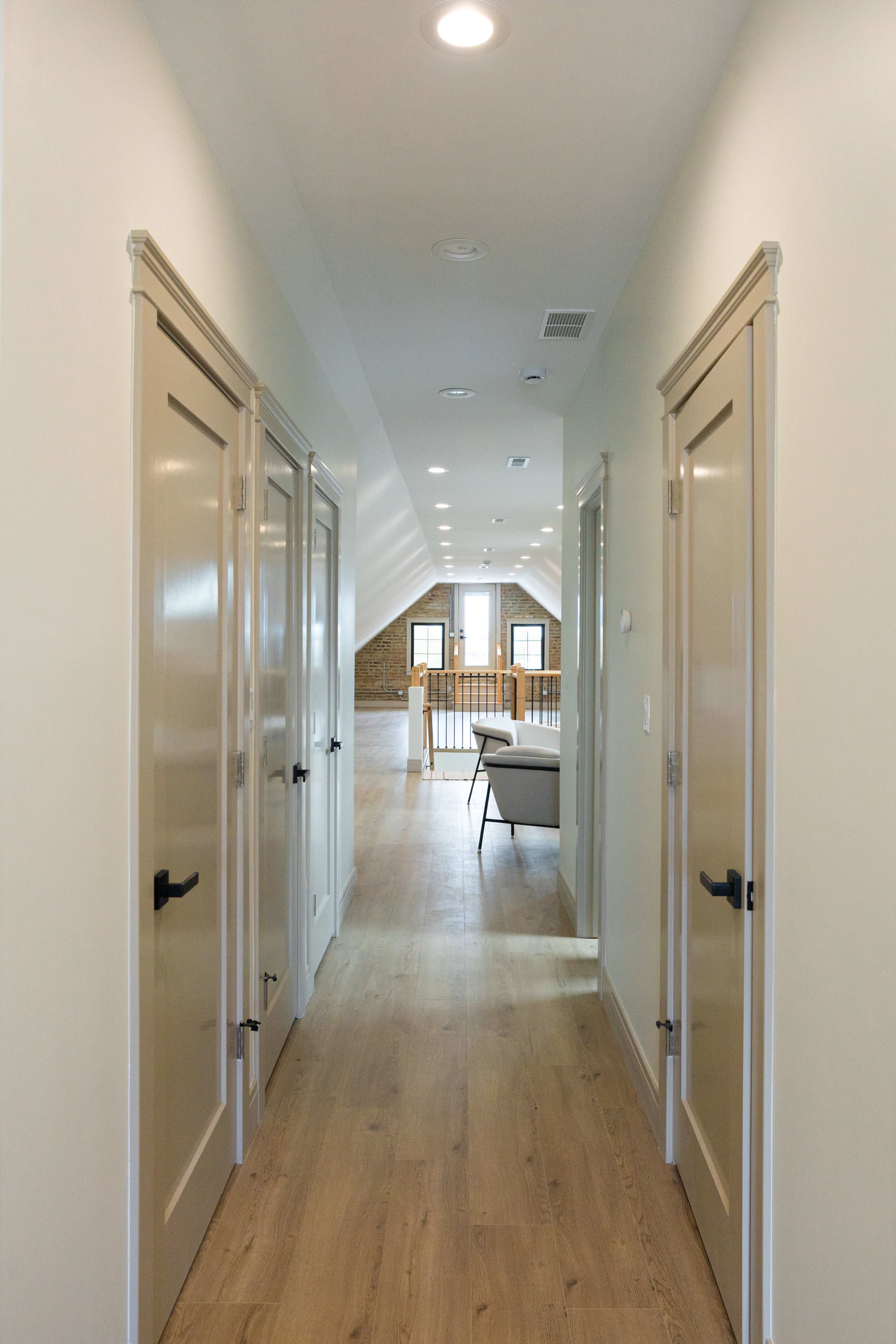 The image shows a long, well-lit hallway with several closed doors on either side, leading to a spacious room at the end.