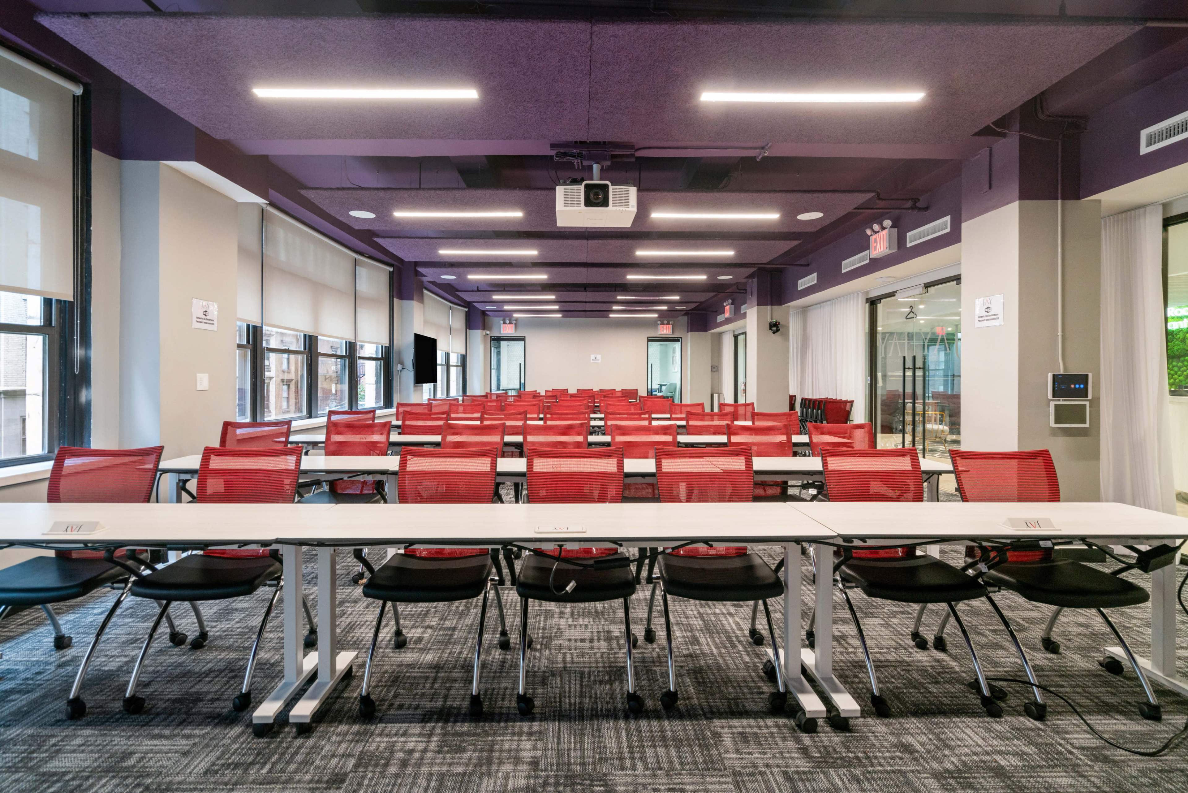 A conference room features rows of red chairs facing a long white table, with a projector mounted on the ceiling and large windows allowing natural light.