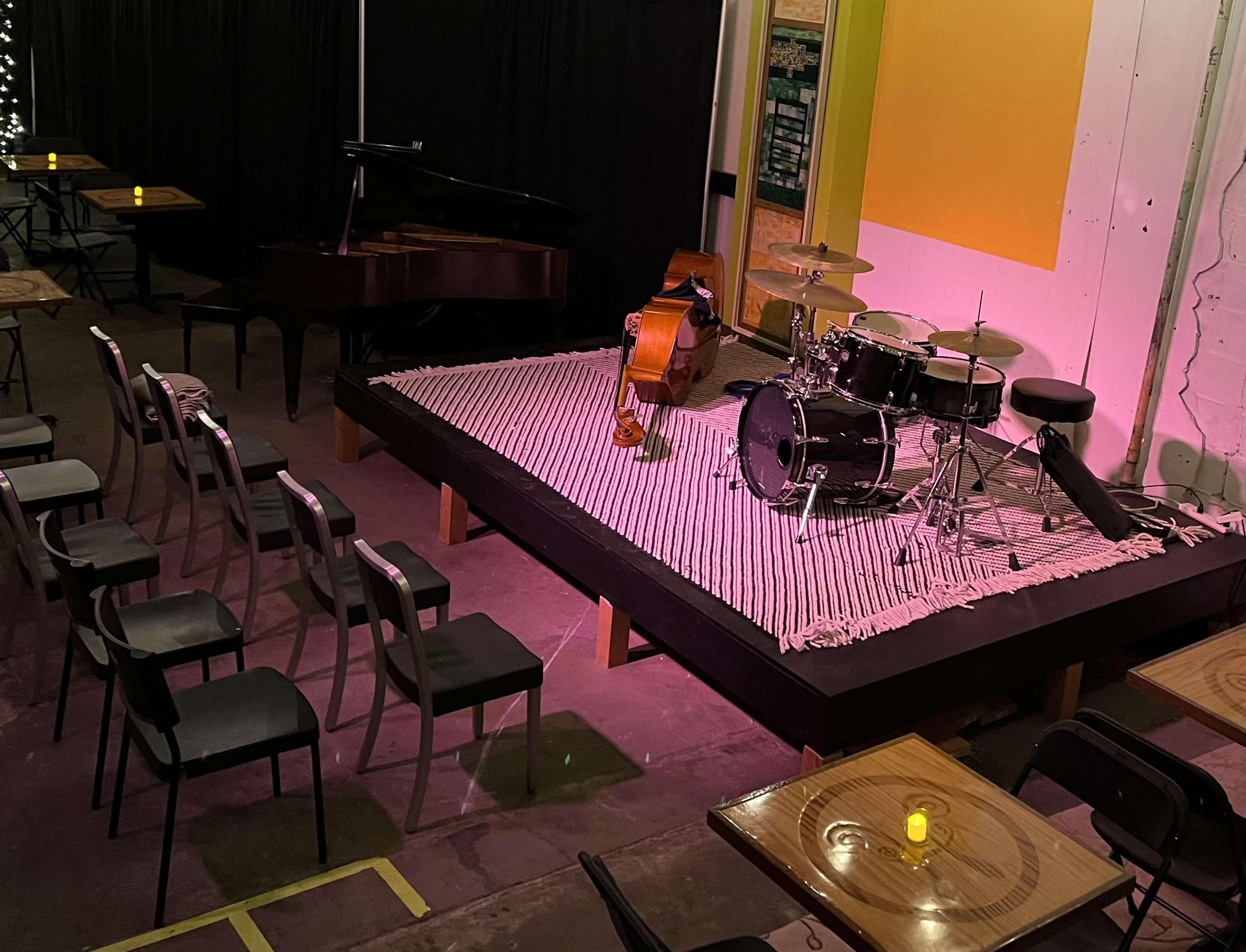 A small performance stage with a grand piano, double bass, drum set, and several rows of empty chairs arranged around tables.