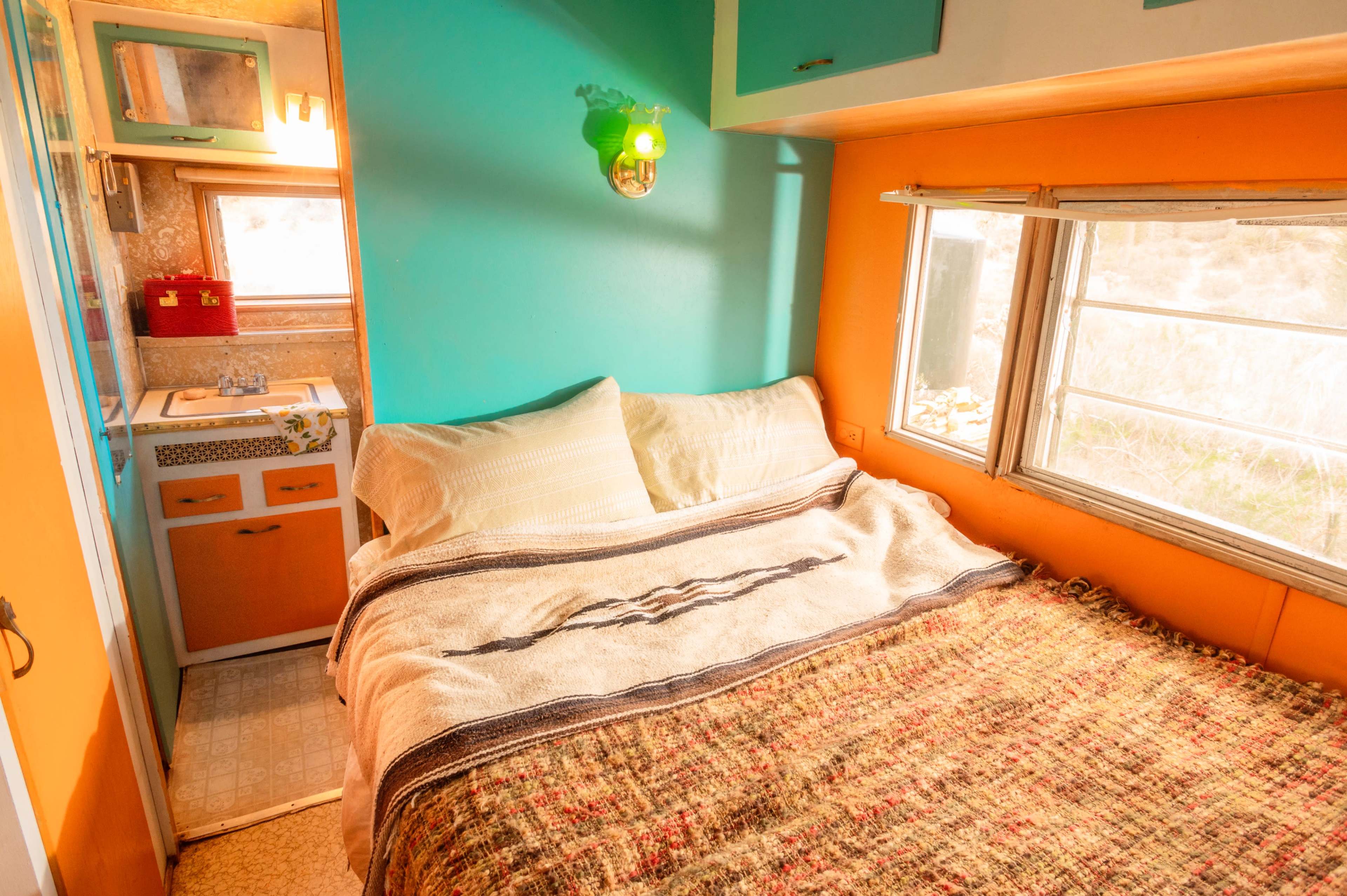 A cozy interior of a small trailer with a bed covered in a woven blanket, and a kitchenette visible in the background.