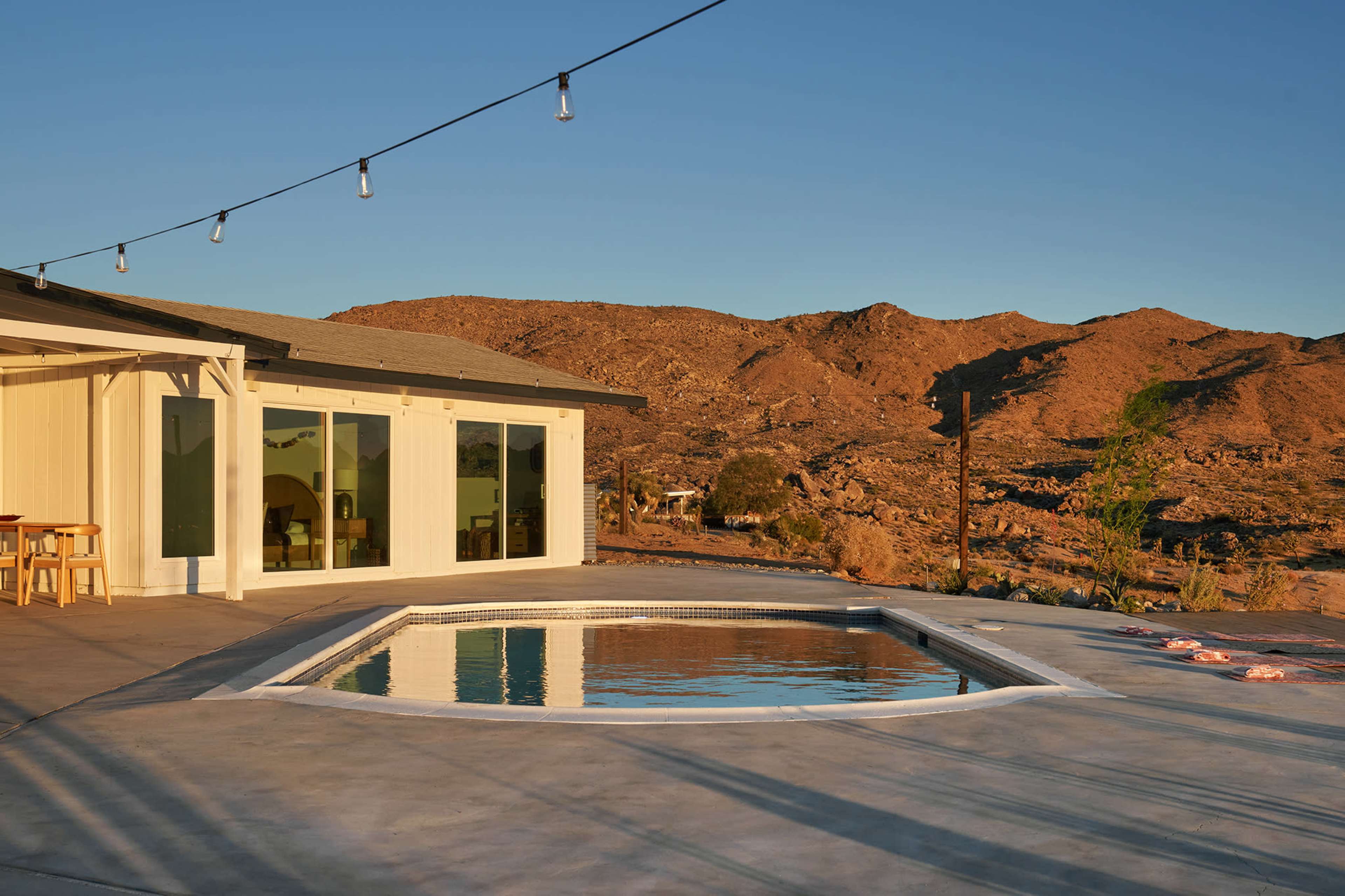 A modern poolside area features a rectangular swimming pool next to a contemporary house, with mountains visible in the background under a clear sky.