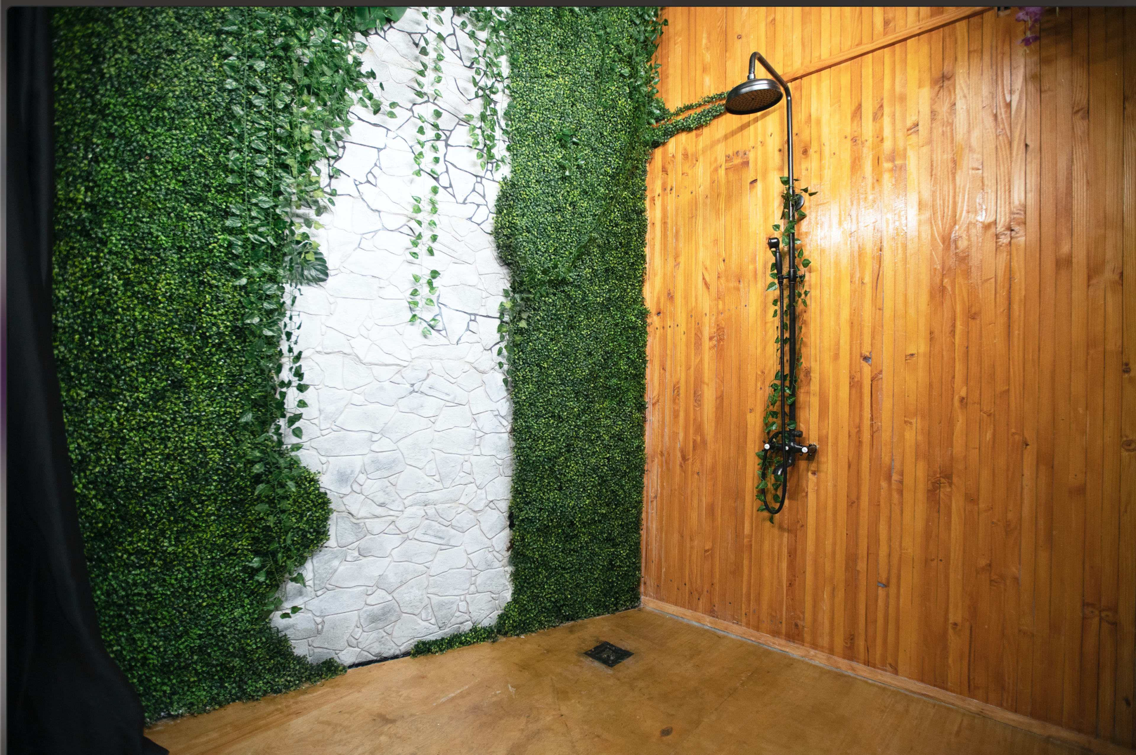 The image features a shower area with a wooden wall on one side and a vertical arrangement of greenery and a stone-like panel on the opposite side.