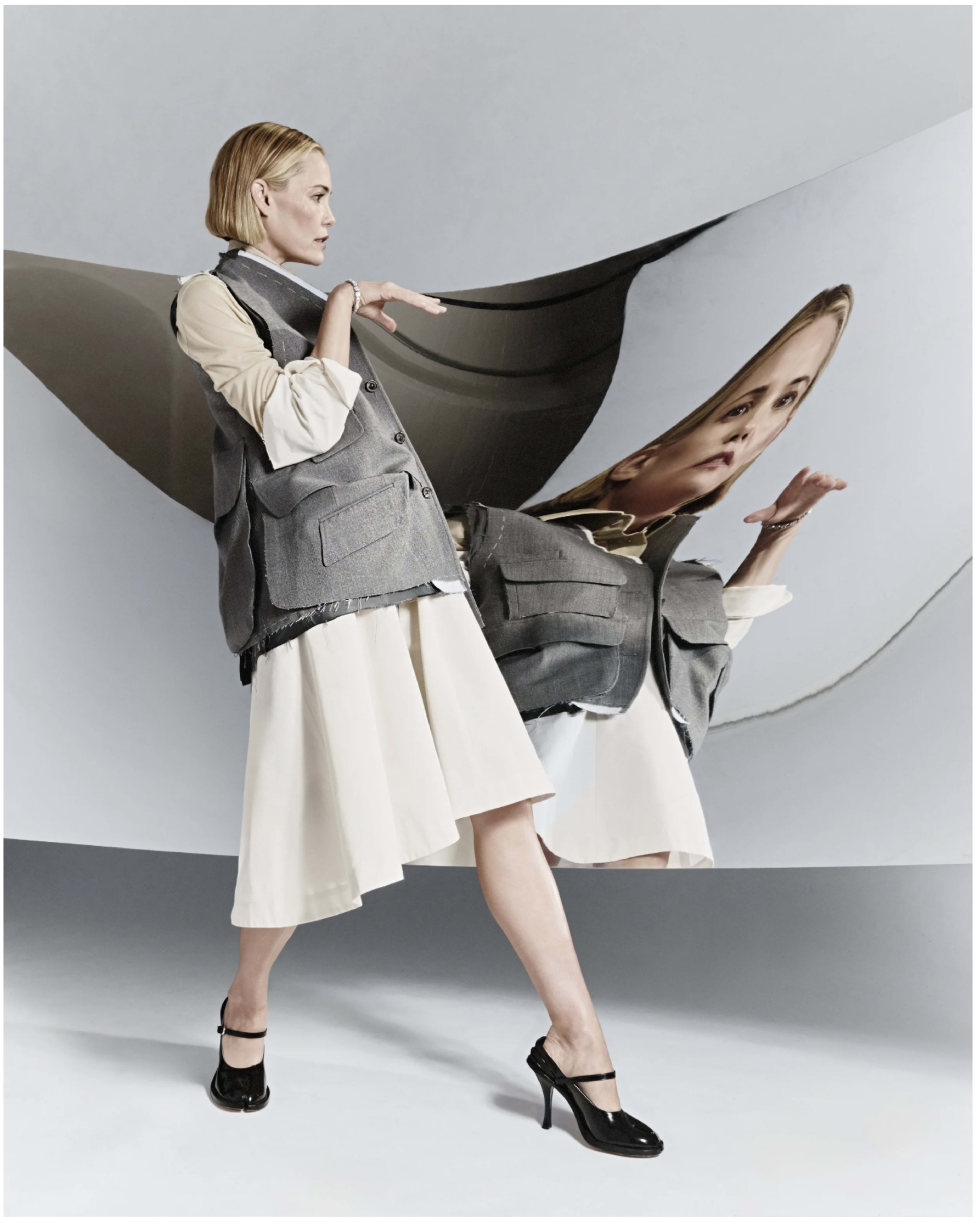 A woman in a gray vest and white skirt stands in front of a reflective, wavy surface that distorts her image.