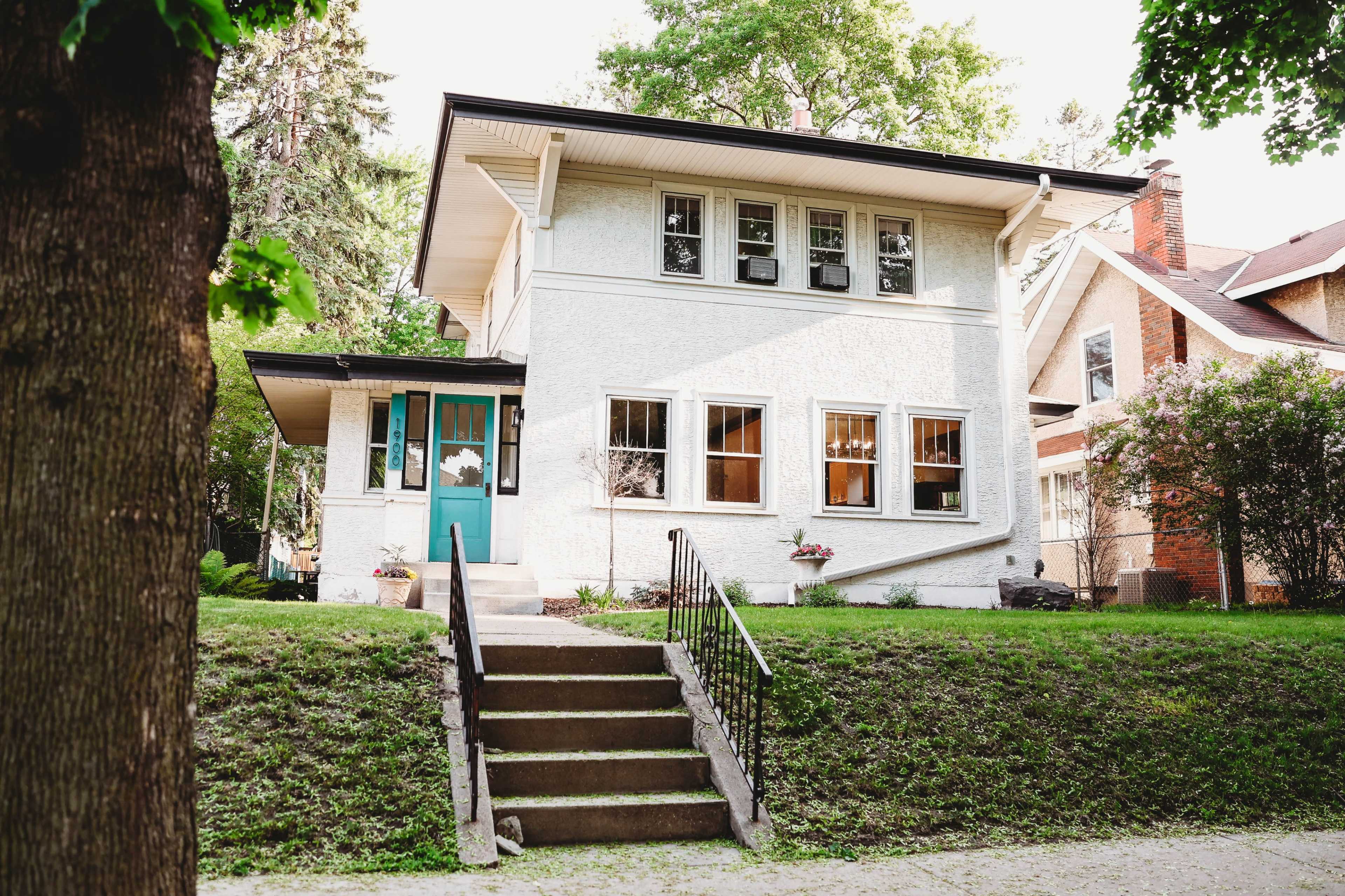 A two-story white house with a turquoise door and several windows is situated on a grassy lawn beside a tree-lined street.