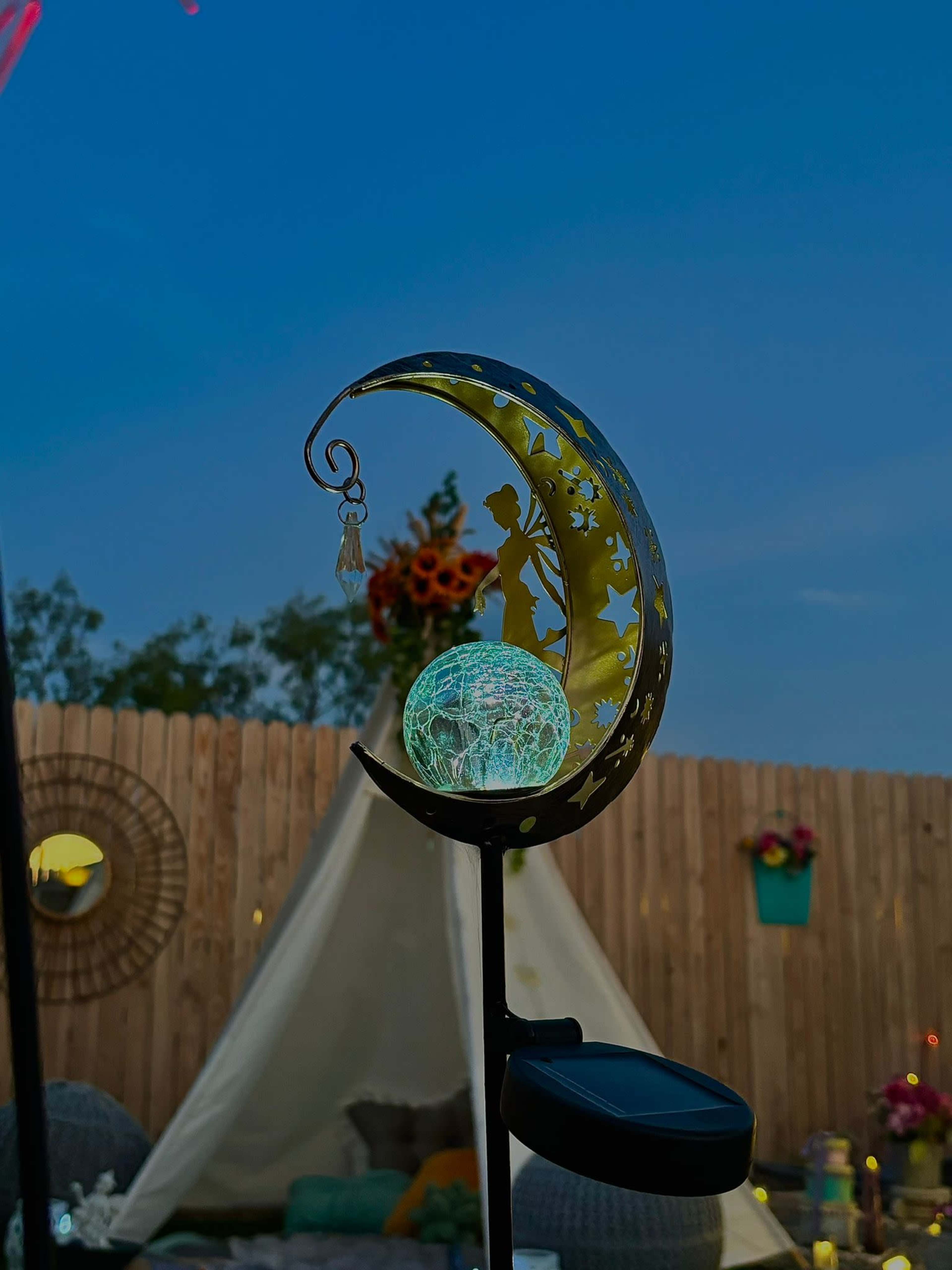 A crescent moon decoration with a fairy figure holds a glowing orb in front of a tent and wooden fence.