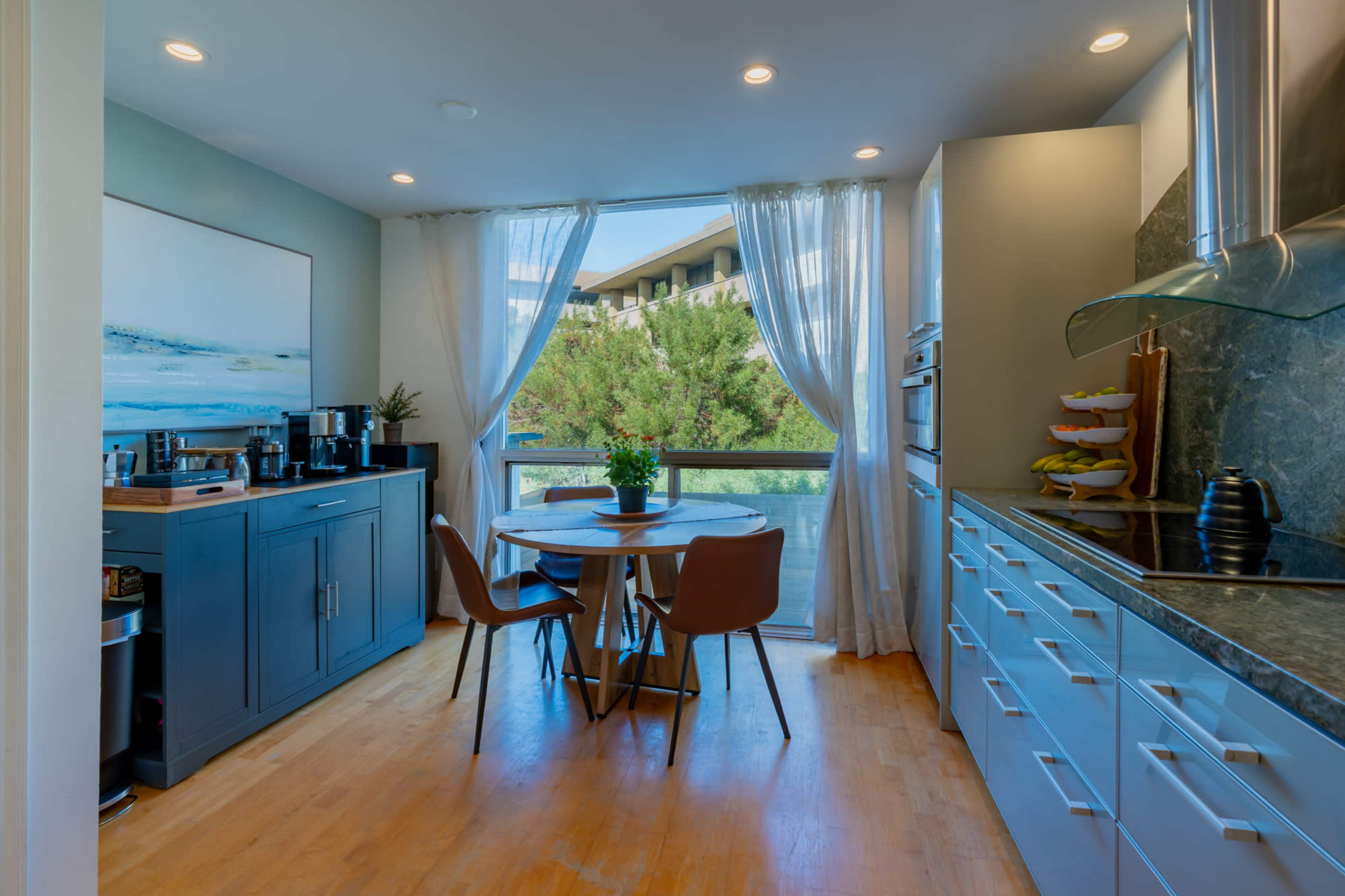 A modern kitchen features a circular wooden table surrounded by chairs, with a large window allowing natural light and a view of greenery outside.
