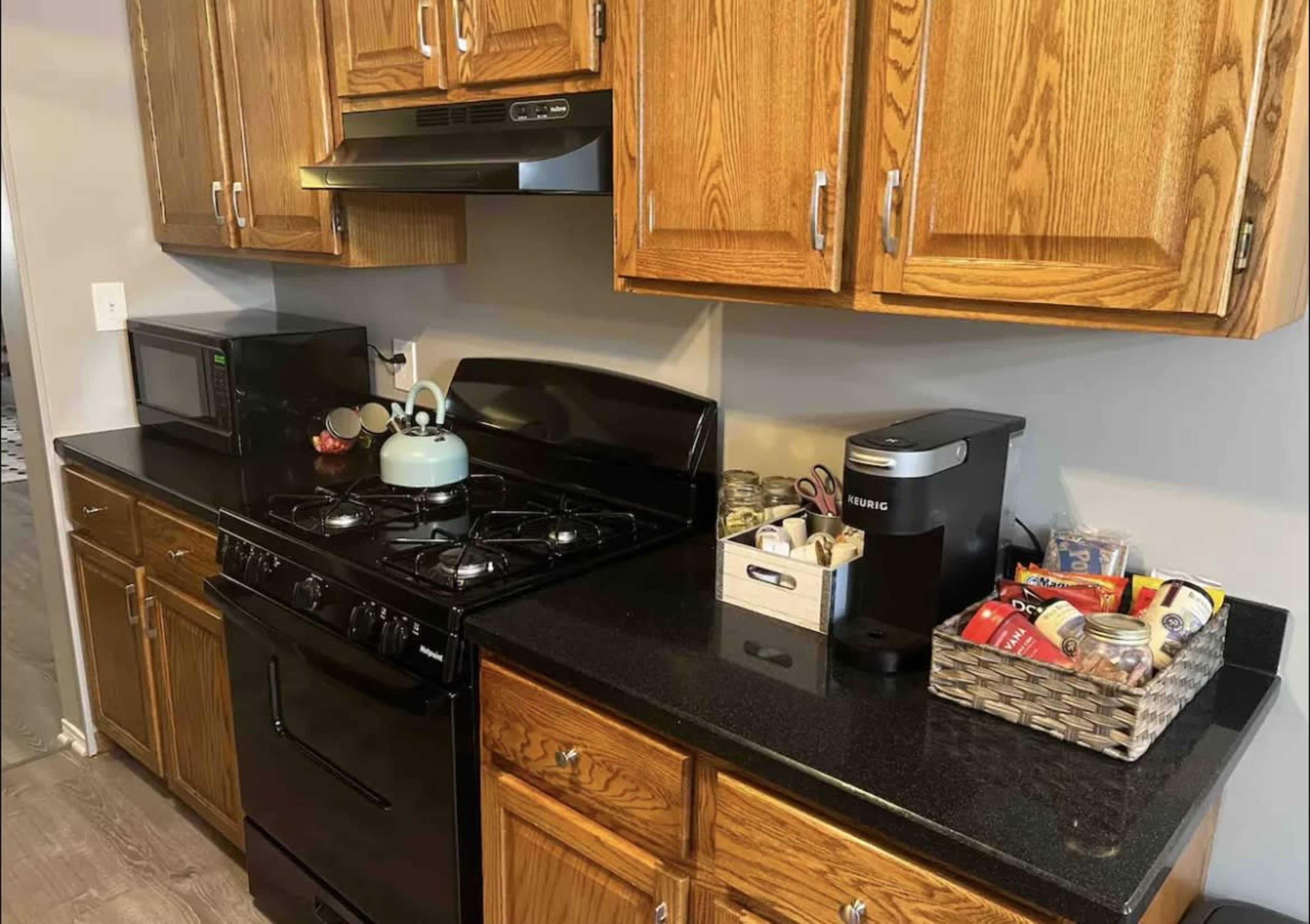 A kitchen counter with a black gas stove, a blue kettle, a microwave, and a coffee maker, surrounded by wooden cabinets and a basket containing various food items.