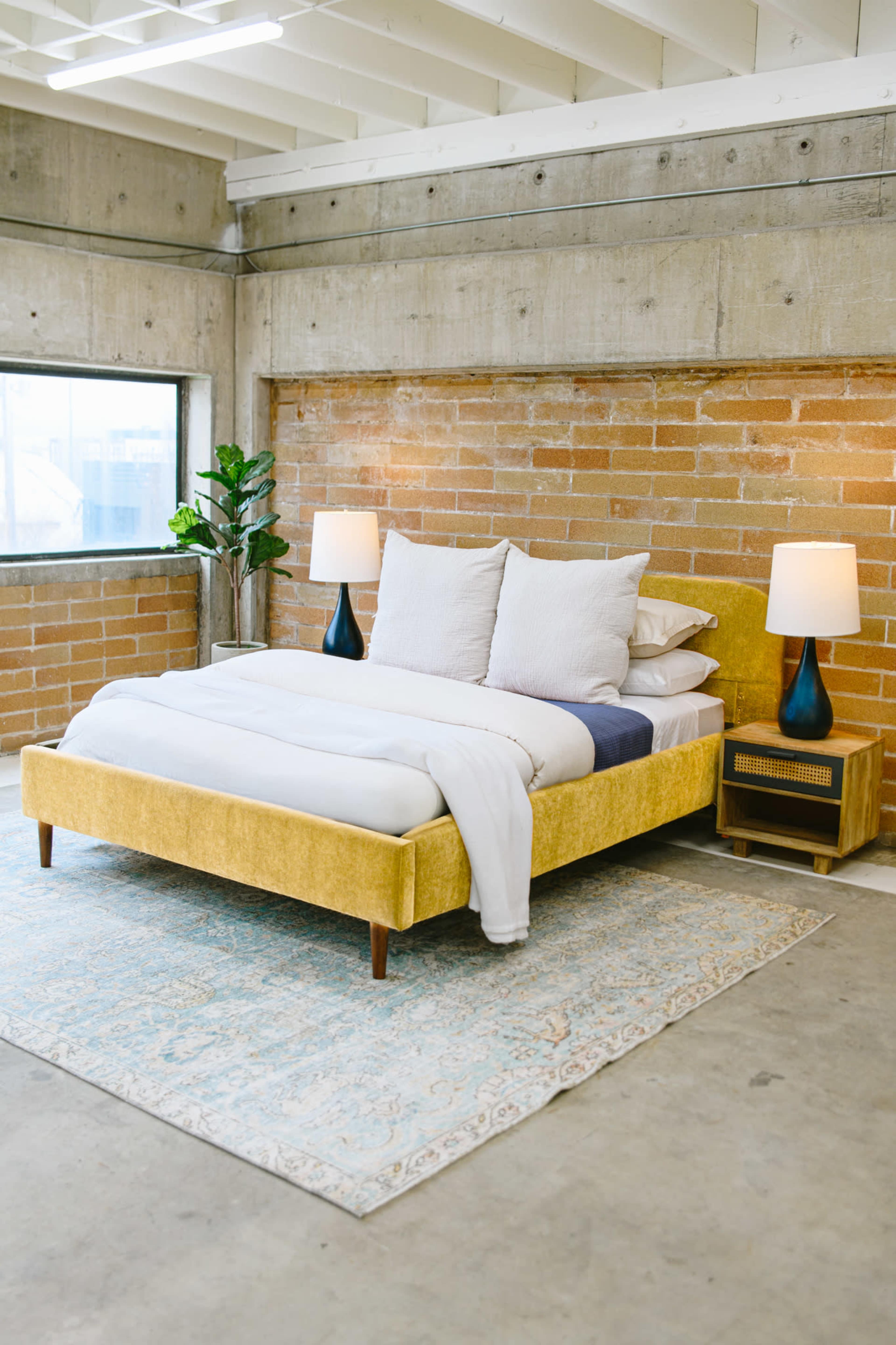 A yellow upholstered bed with white bedding is positioned against a brick wall, accompanied by two blue table lamps and a patterned rug on a concrete floor.