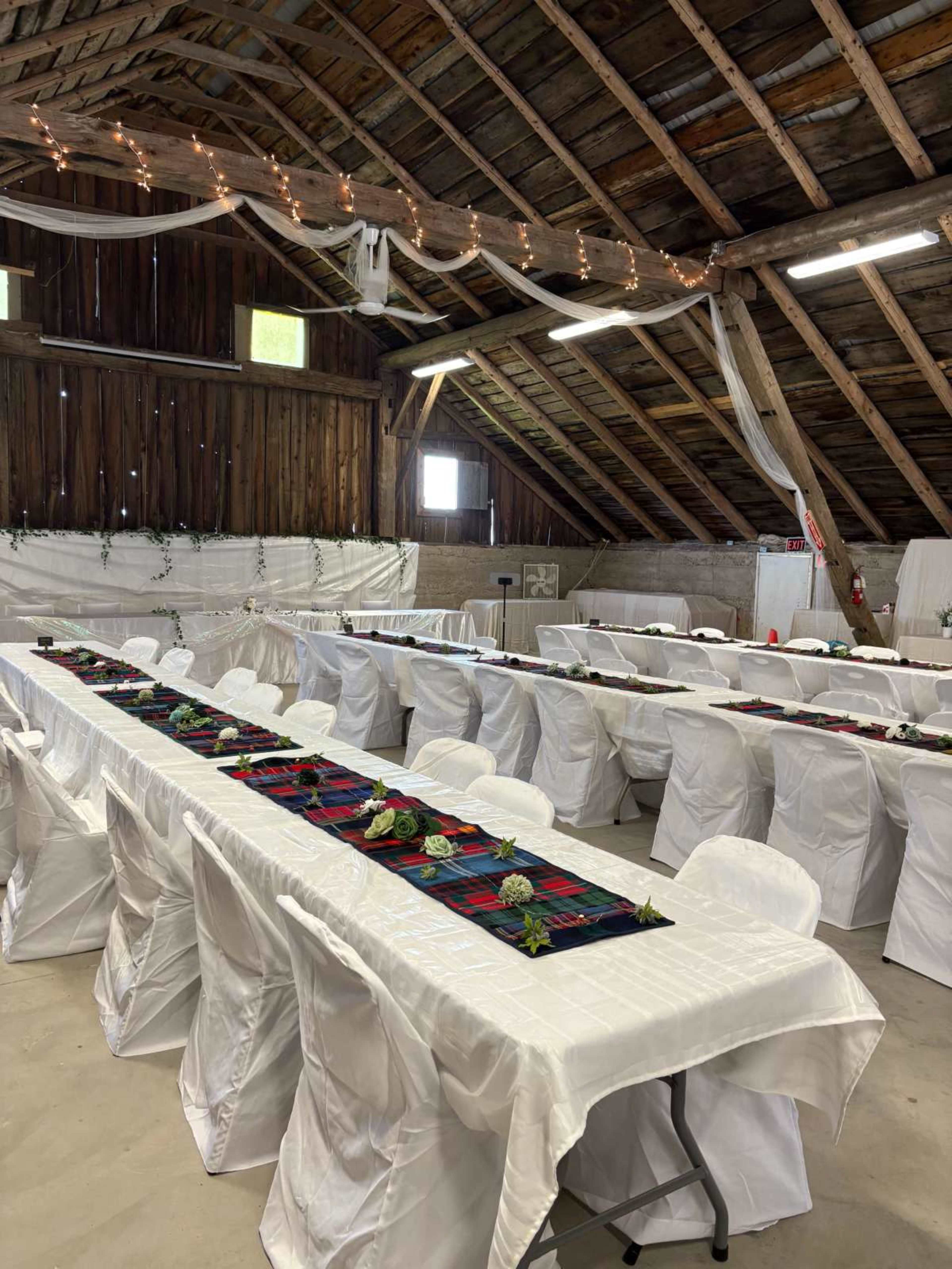 The interior of a rustic barn features rows of long tables covered with white cloths and adorned with floral centerpieces, illuminated by string lights hung from the ceiling.