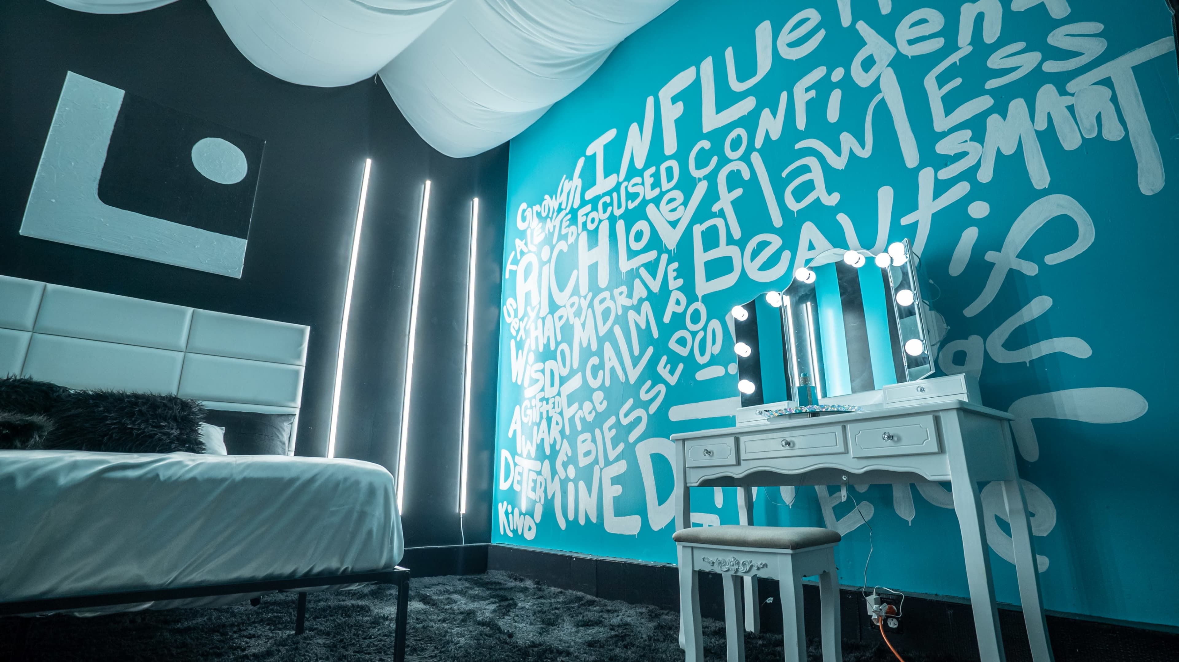 The image shows a modern bedroom with a white bed, a vanity table with mirrors and lights, and a feature wall covered in various inspirational words and phrases.