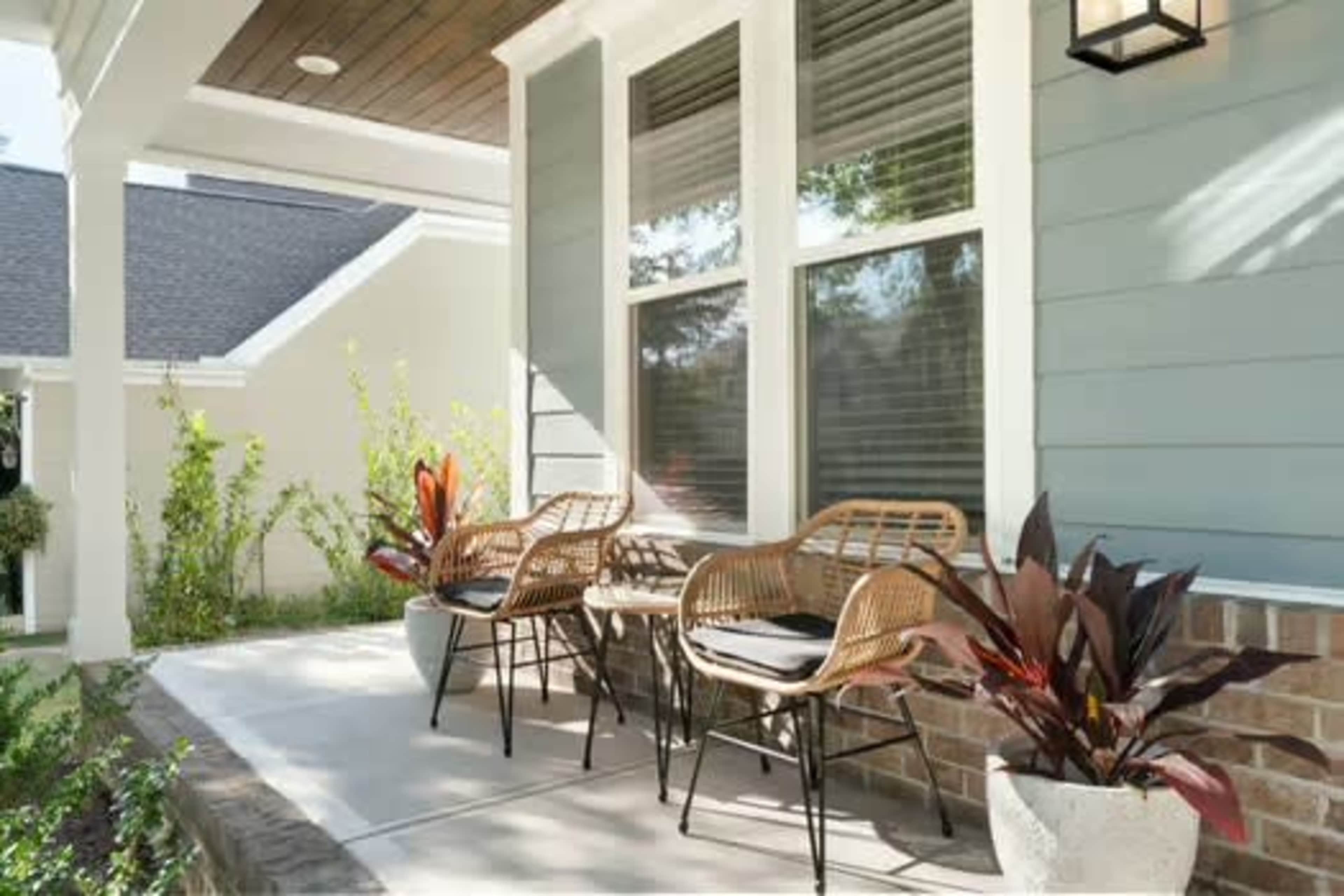 A porch features two wicker chairs and potted plants, framed by large windows on a light blue house.