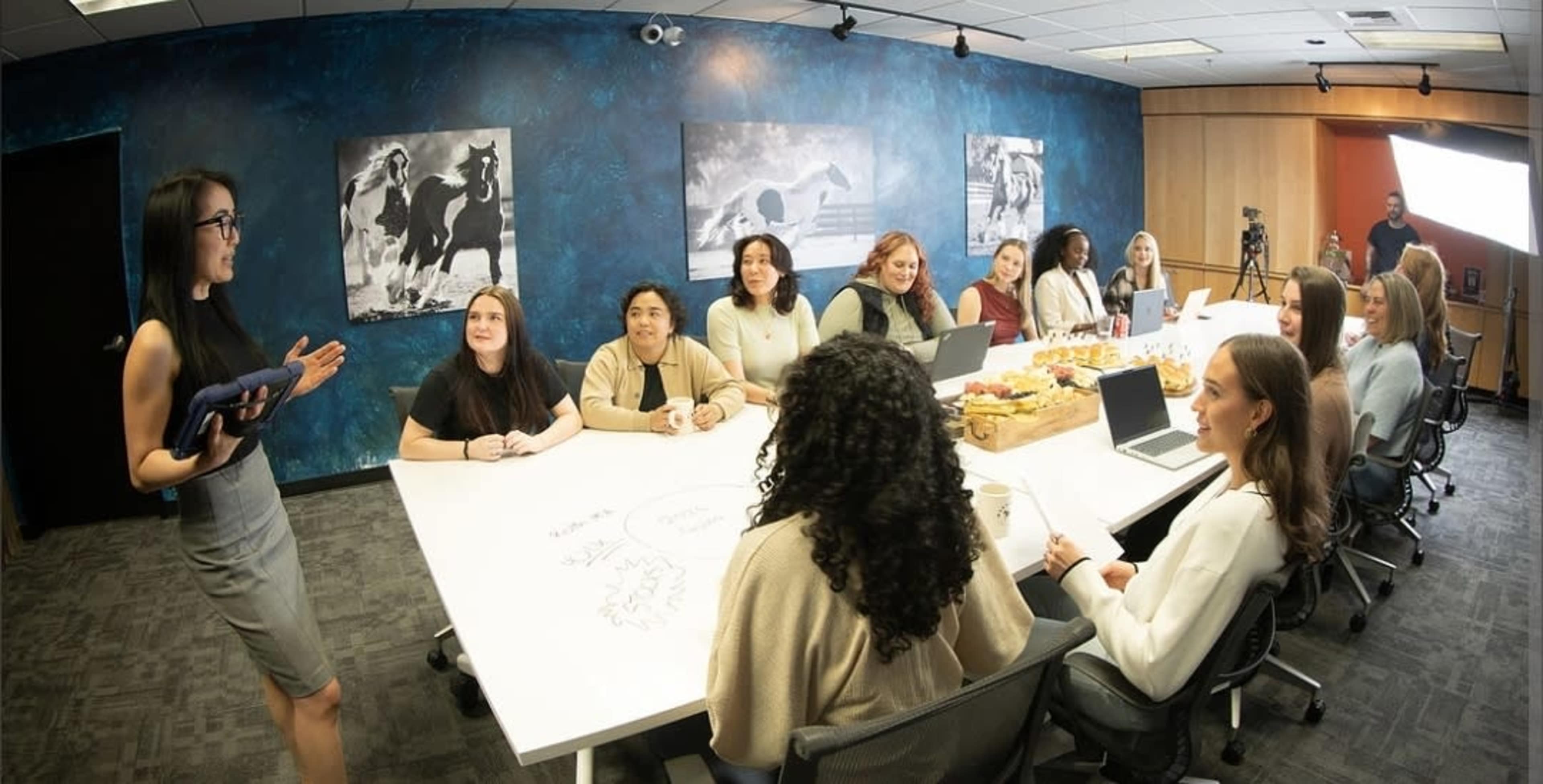 A group of participants sits around a large conference table in a modern meeting room while a presenter stands and addresses them.