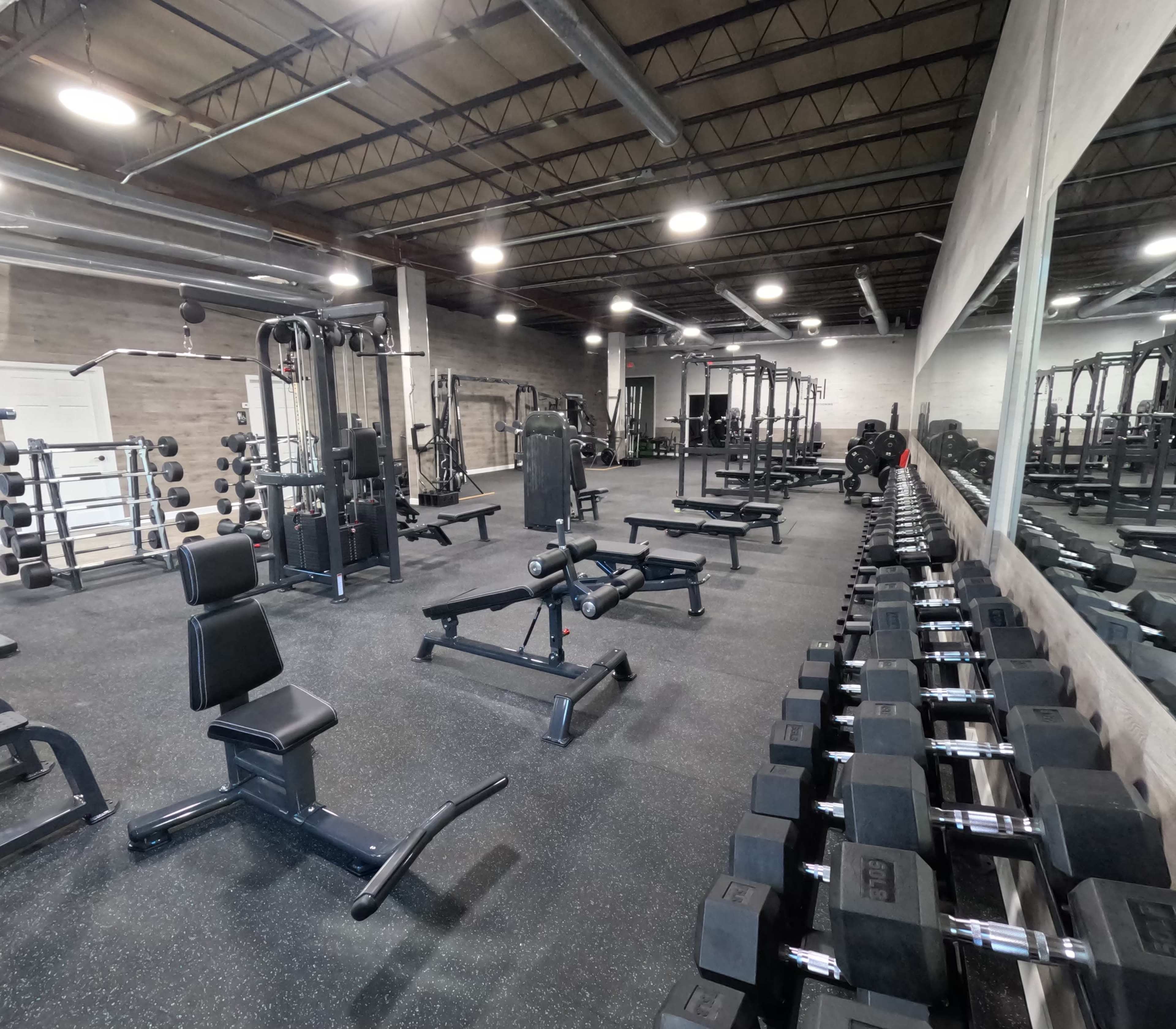 A spacious gym interior with various weightlifting equipment, including a row of dumbbells and benches, along with weight machines arranged across the room.