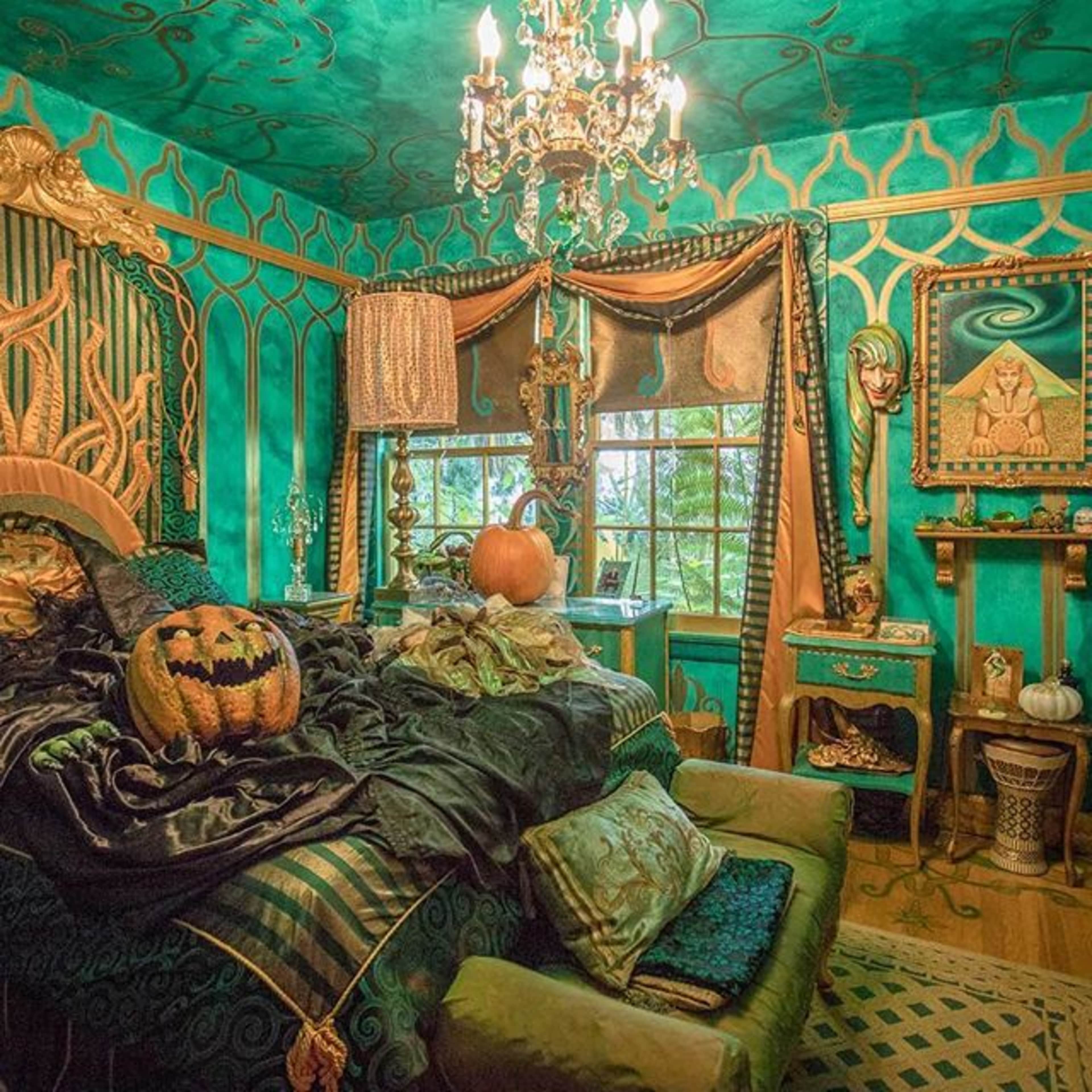 The room features vibrant teal walls, a large bed with dark bedding, a pumpkin on the bed, and ornate decorations including a chandelier and framed artwork.