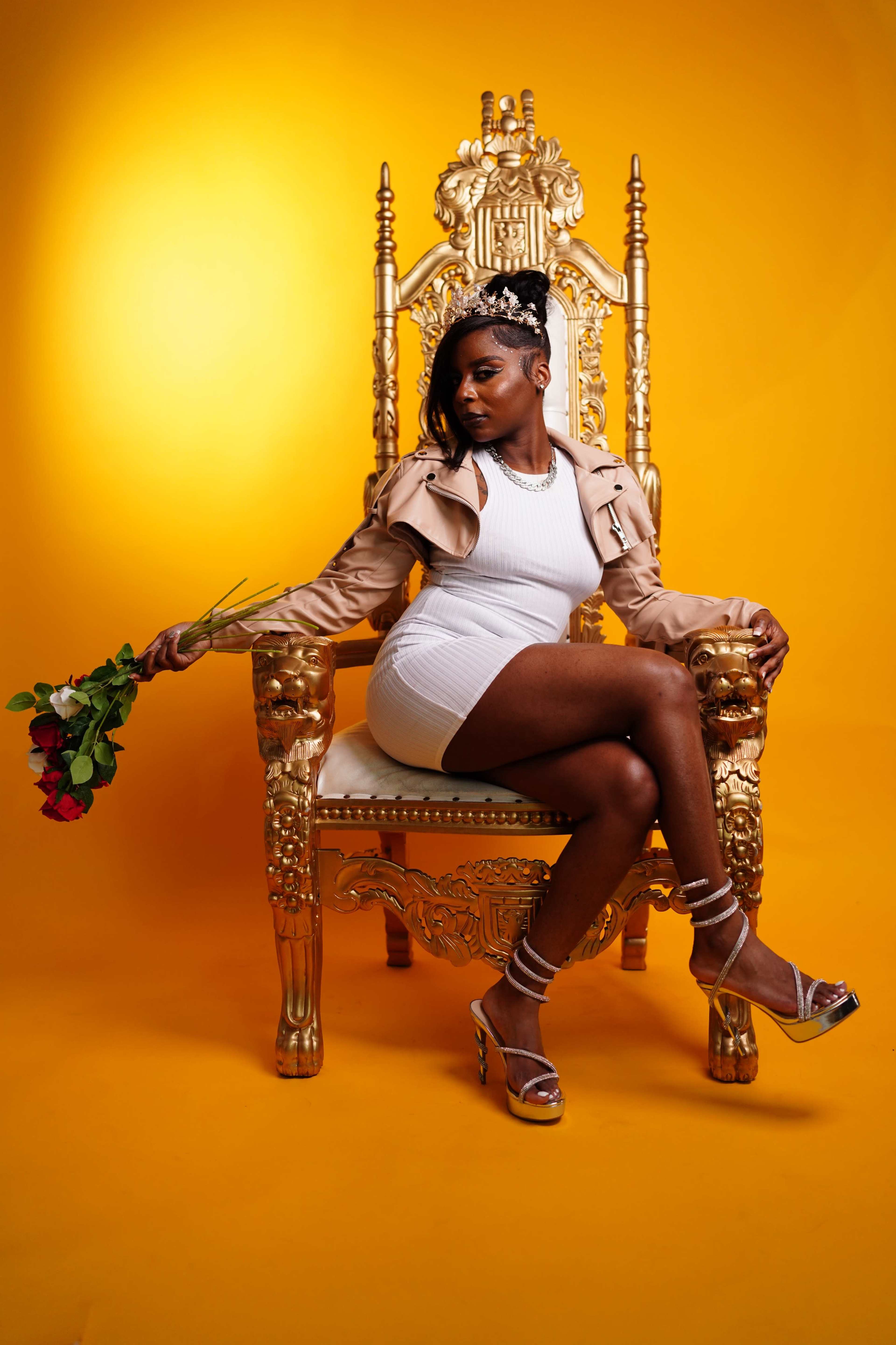 A person sits on an ornate golden throne against a vibrant yellow background, holding a bouquet of red roses.