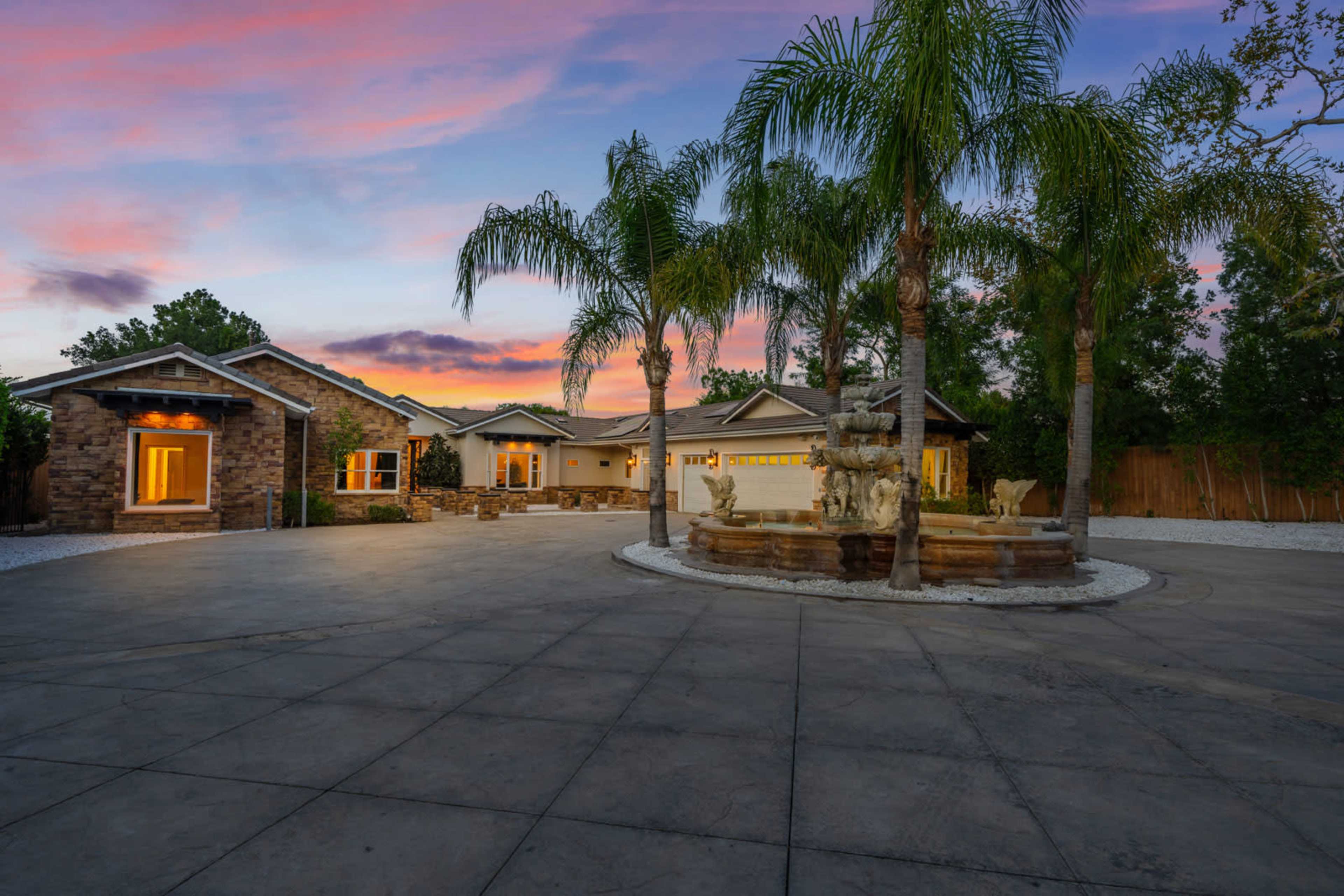 A modern home with a stone facade and landscaped front yard features a circular driveway and a fountain surrounded by palm trees at sunset.