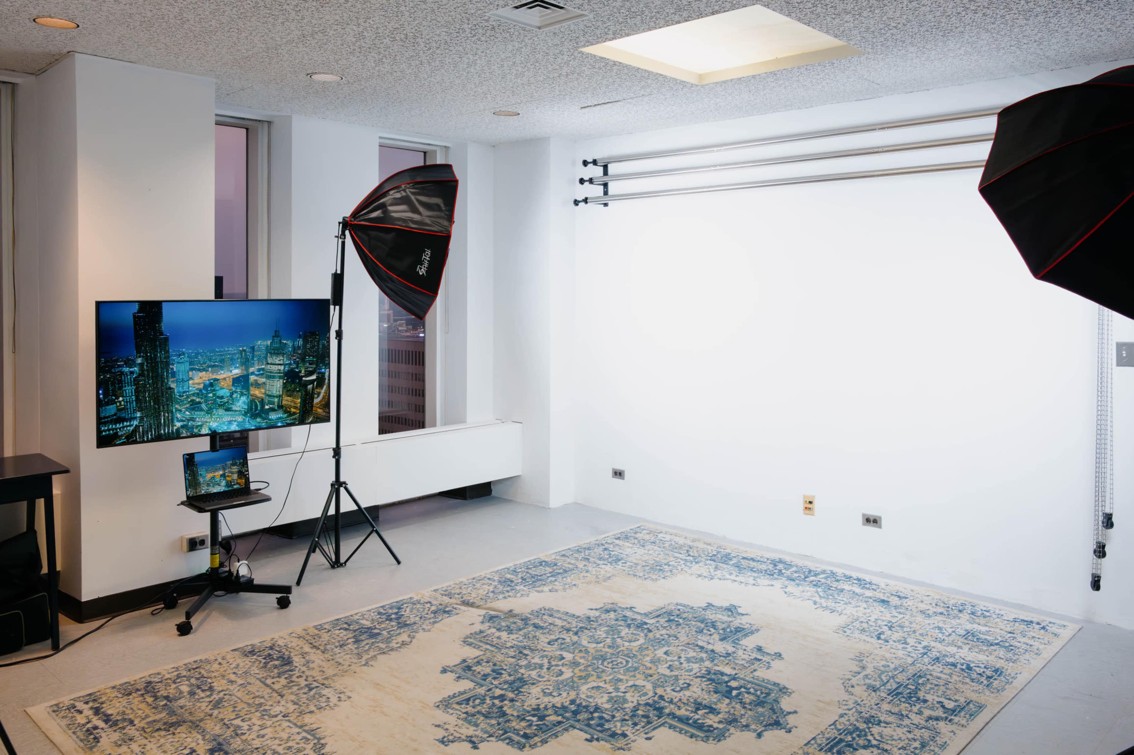 The image shows a photography studio setup featuring a blank white backdrop, two softbox lights, and a monitor displaying a cityscape, all arranged on a patterned rug.