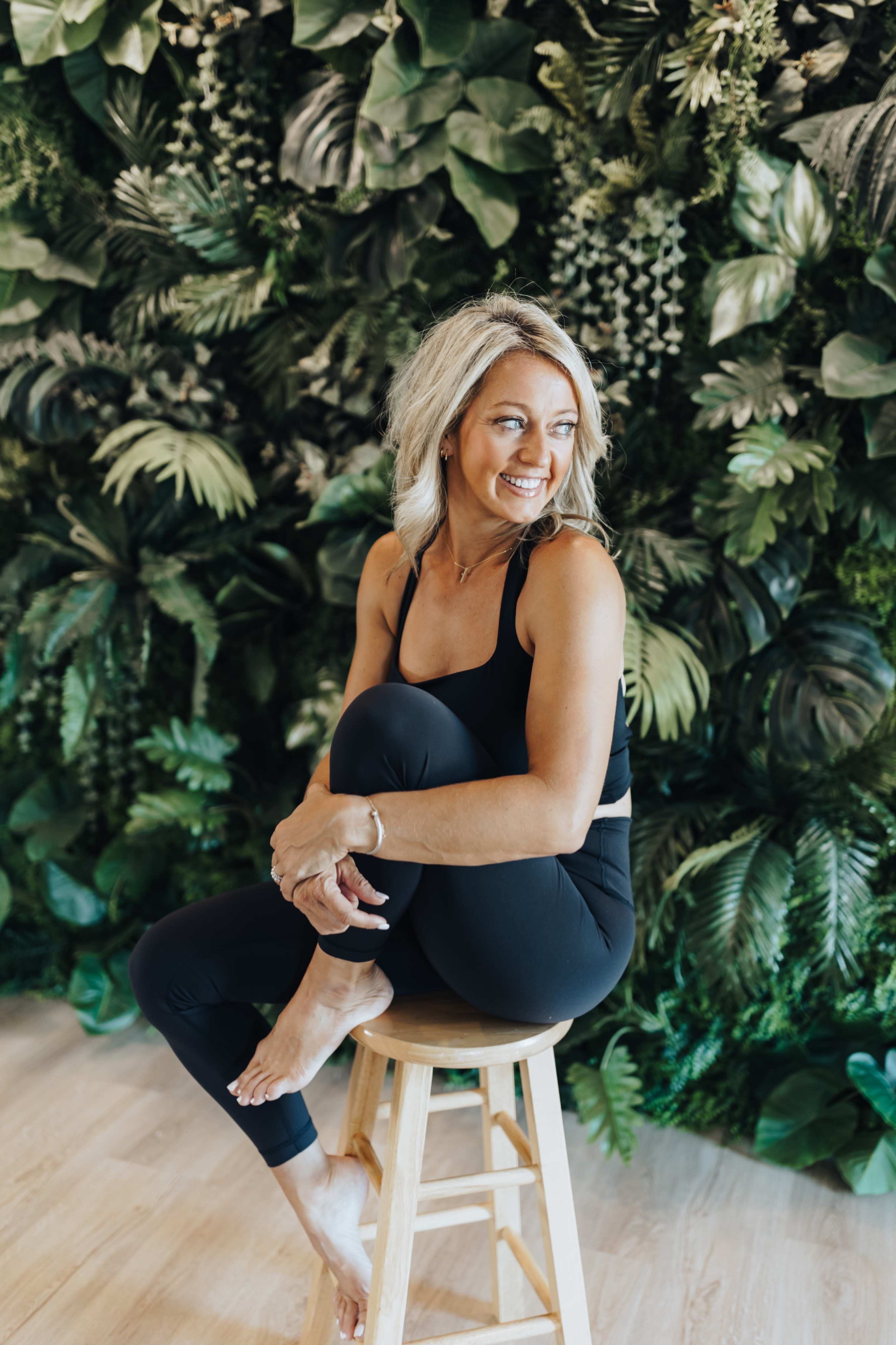 A woman in black athletic wear sits on a wooden stool in front of a lush green plant backdrop.