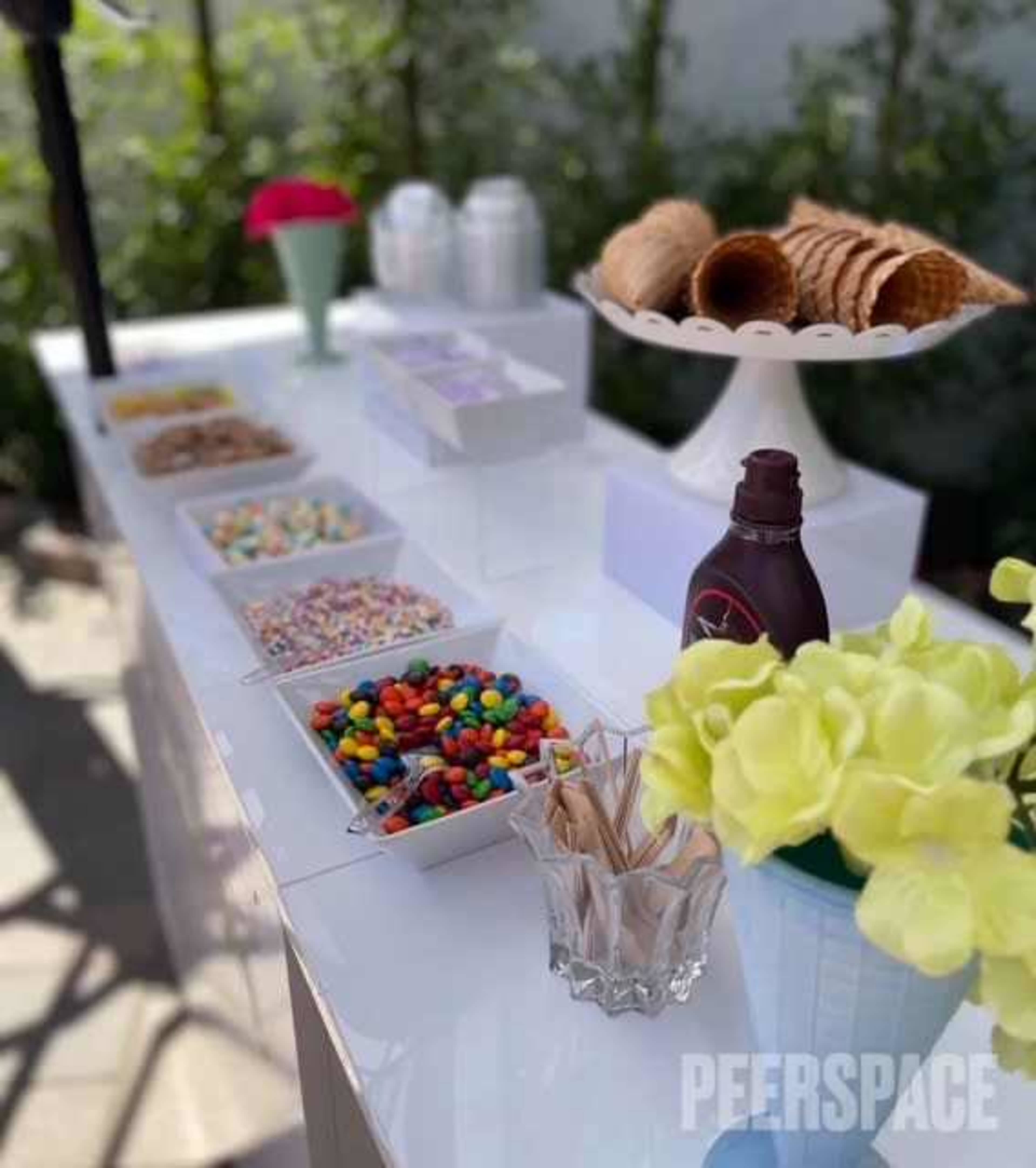 The image shows a candy station featuring various containers of colorful candies, sprinkles, and waffle cones, arranged on a white table with decorative flowers.