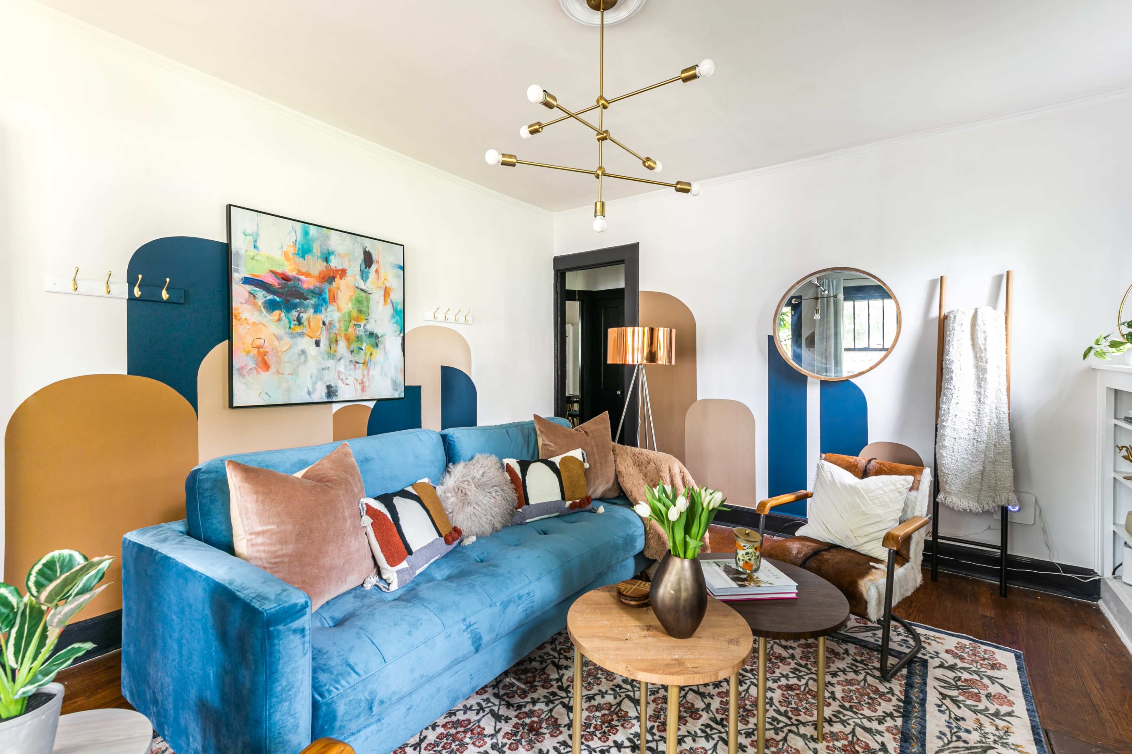 A living room featuring a blue sofa with various cushions, a round mirror, a floor lamp, and colorful wall accents.