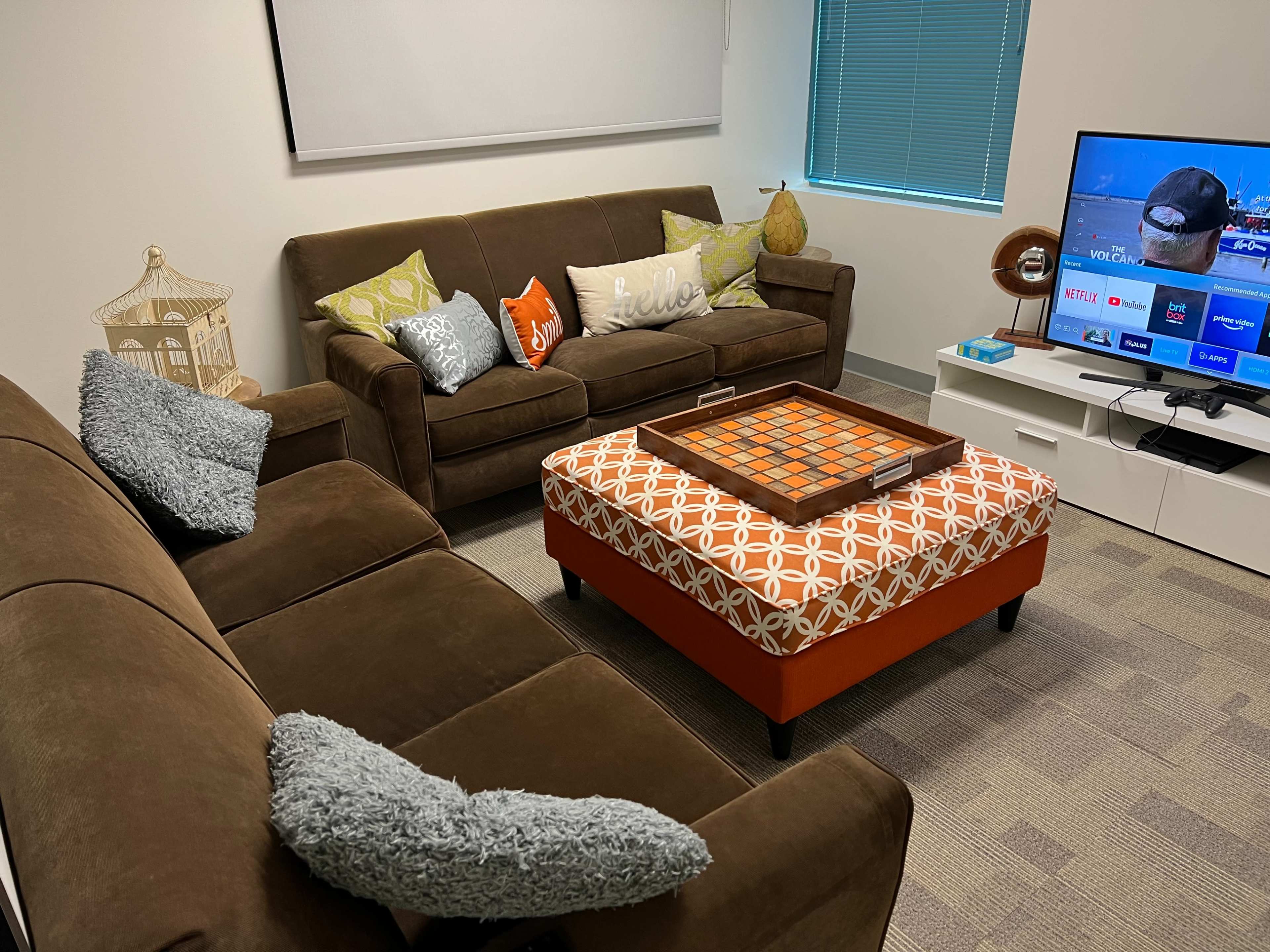 The living room features two brown sofas, a patterned ottoman, a television displaying streaming options, and a window with blinds.
