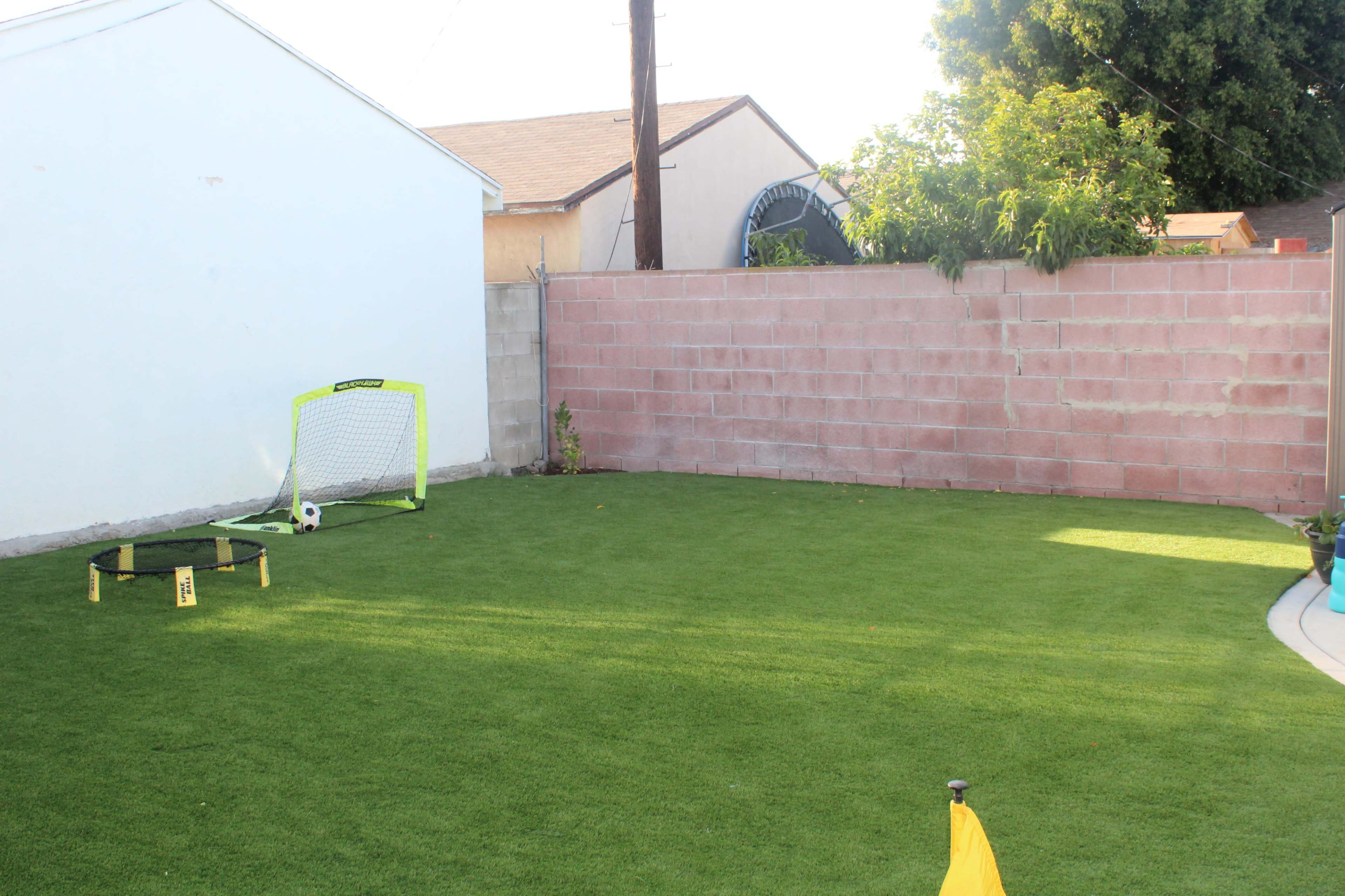 A backyard with artificial grass featuring a small soccer goal and a practice rebounder against a wall.