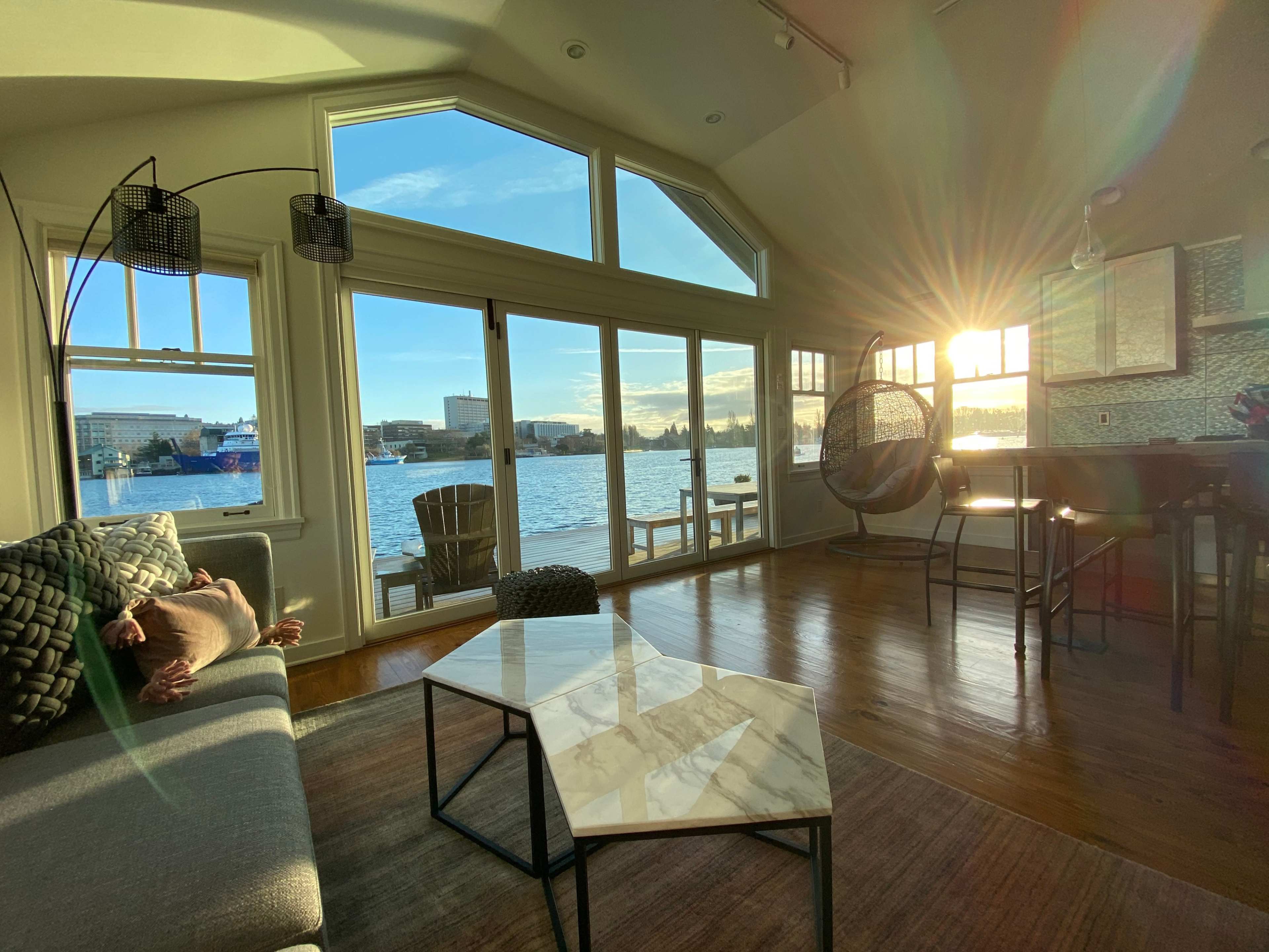 A modern living room features a large window overlooking a body of water, with sunlight streaming in and illuminating the space.