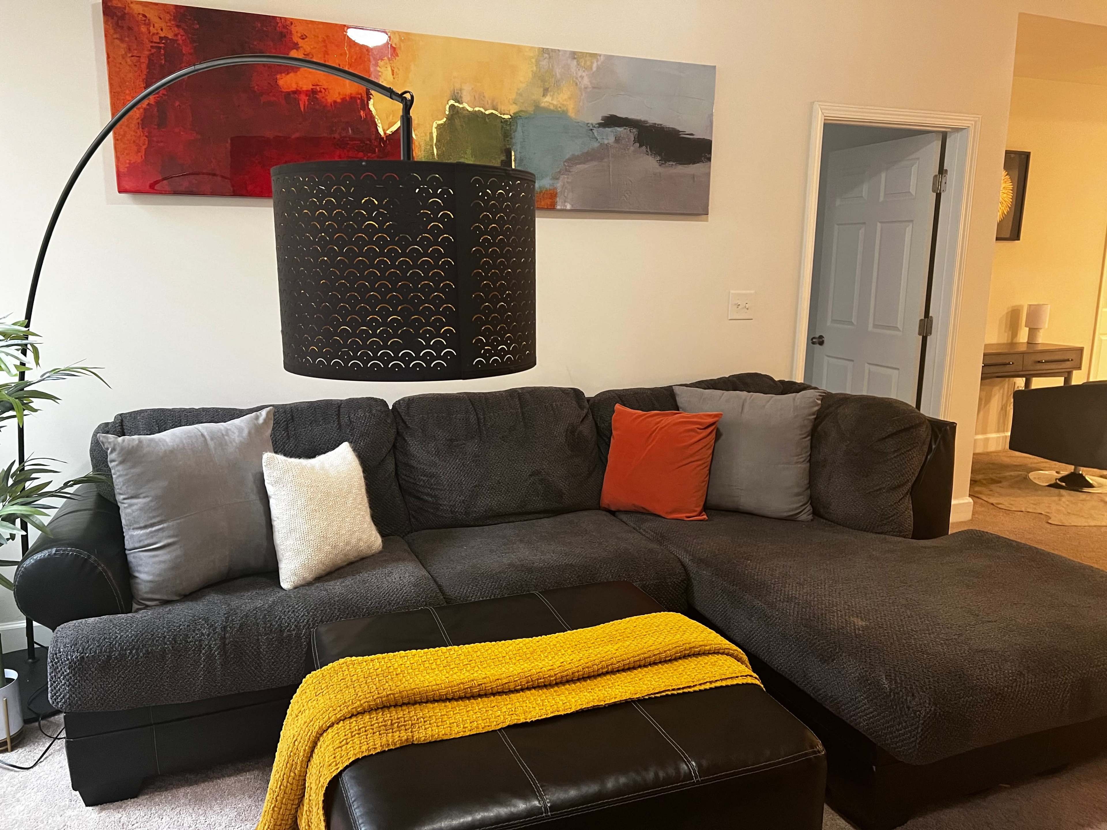 A gray sectional sofa with colorful throw pillows and a yellow blanket is positioned in a living room under a black floor lamp, with abstract artwork on the wall.