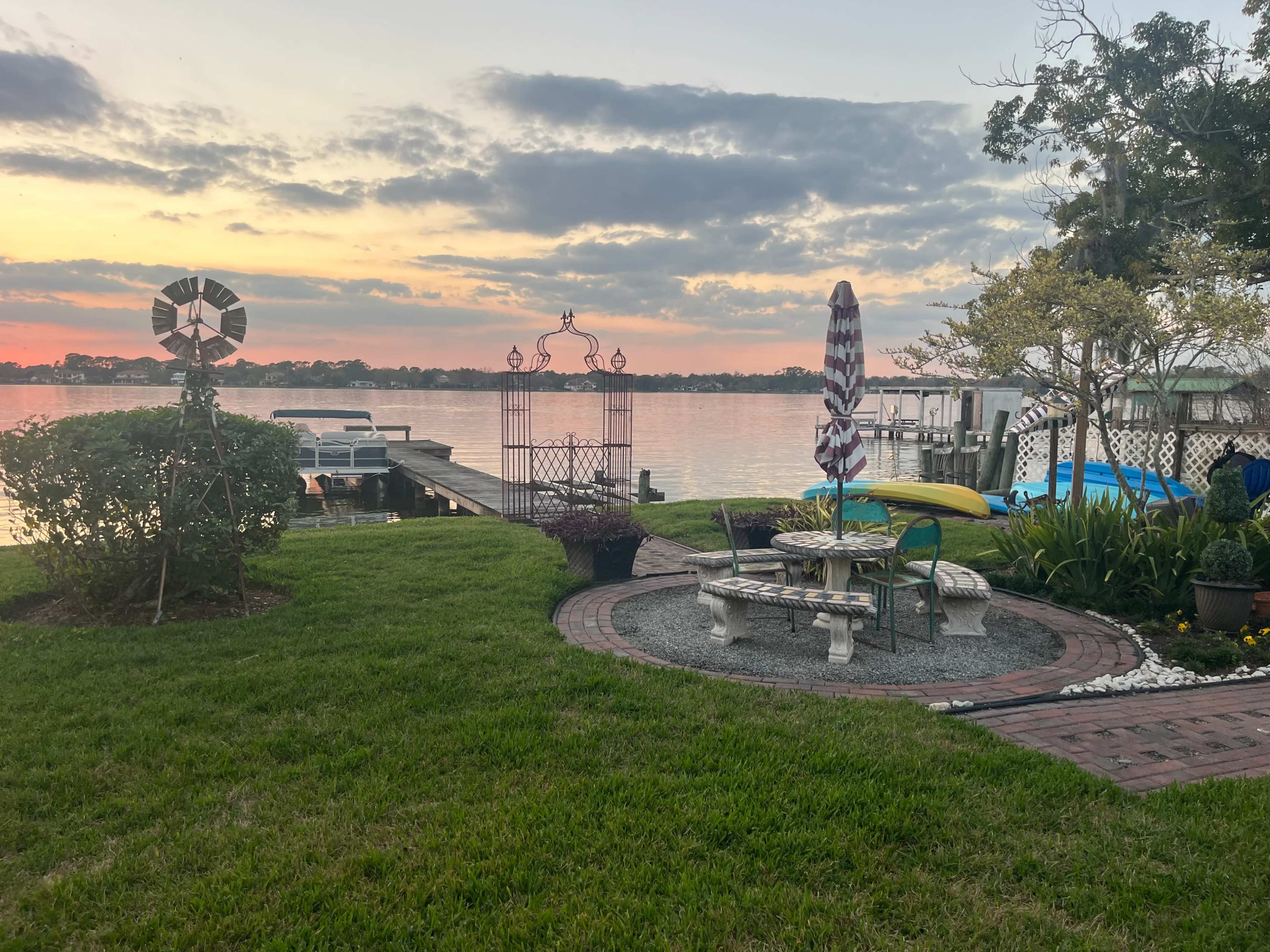 The image shows a lakeside scene with a grassy area, a stone patio with seating, and a decorative gazebo, alongside boats and a dock in the background.