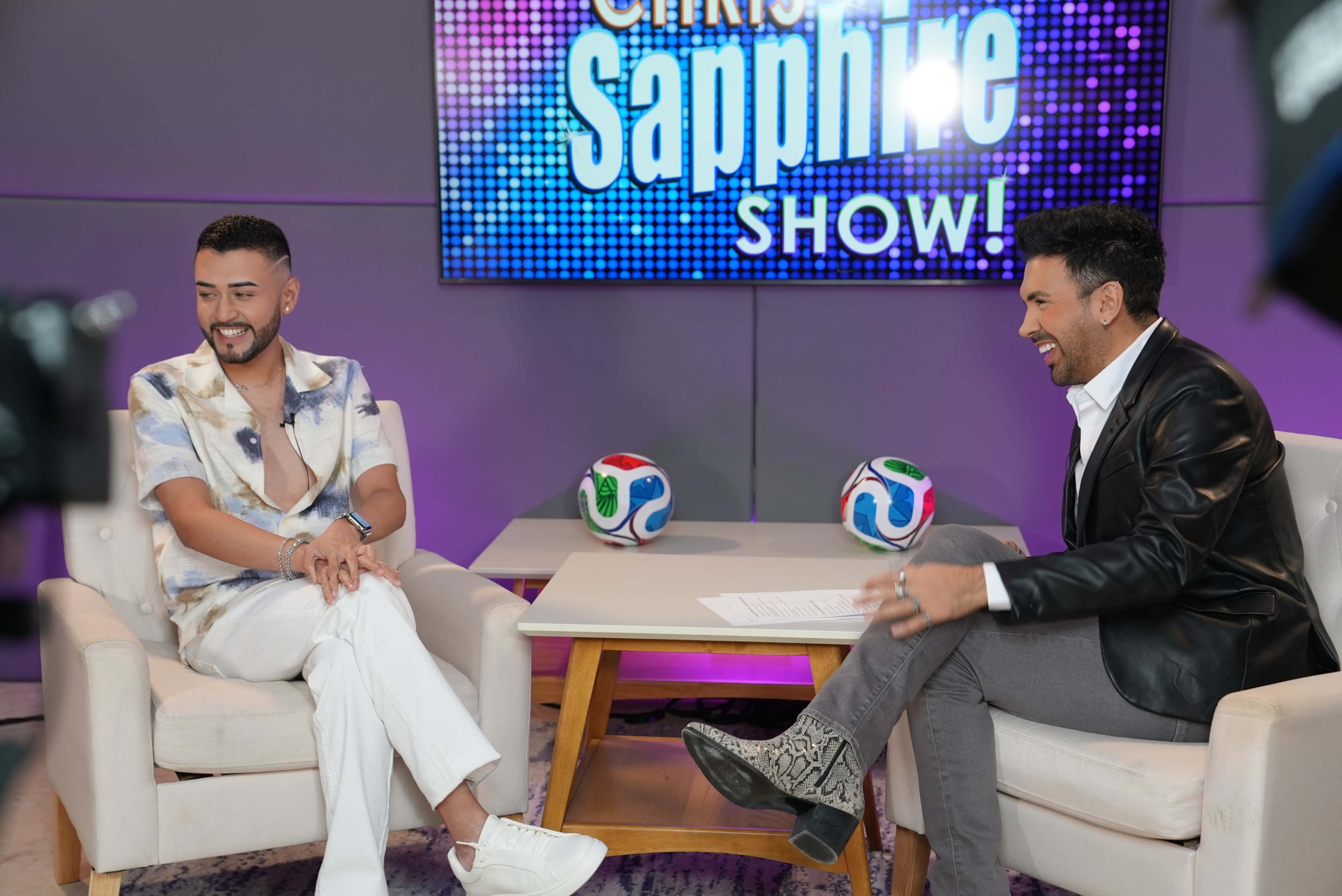Two men are seated on a set, with one wearing a white outfit and the other in a black jacket, as they engage in conversation in front of a colorful backdrop displaying "Sapphire Show."