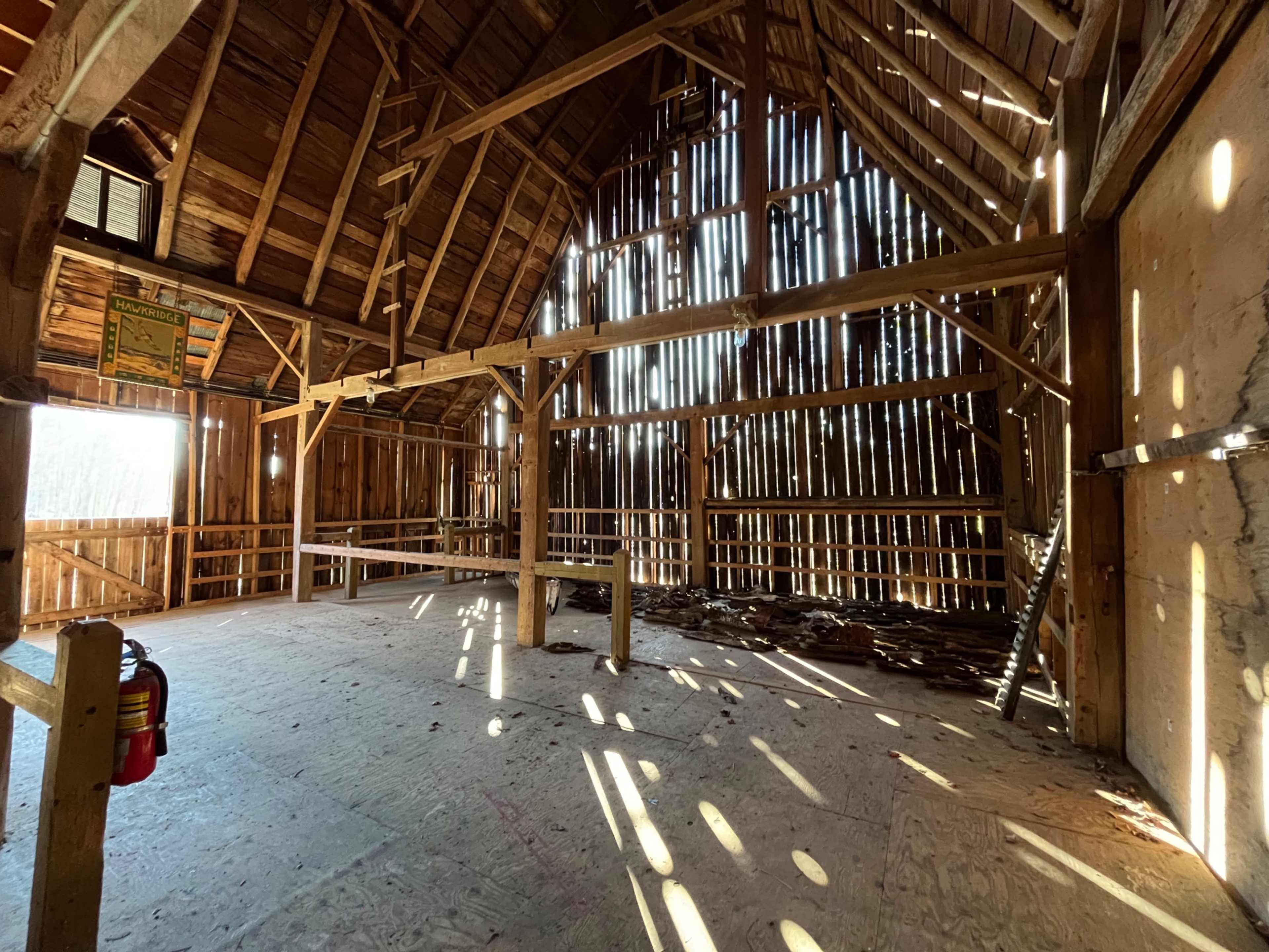 The interior of a rustic barn features wooden beams, a tall ceiling, and sunlight streaming through gaps in the walls.