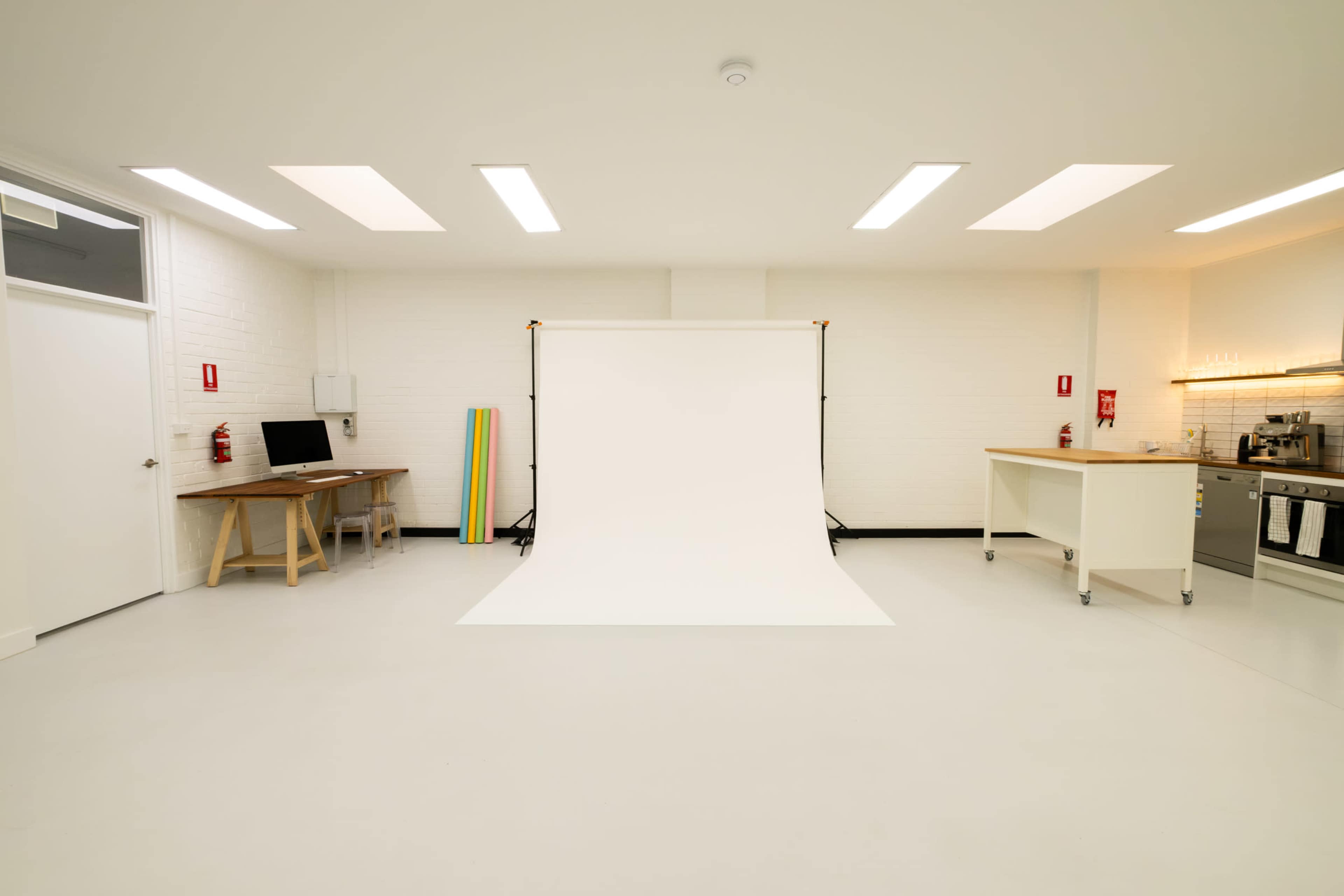 The image shows a minimalist studio space with a white backdrop on a stand, a kitchen area, and various colored props against one wall.
