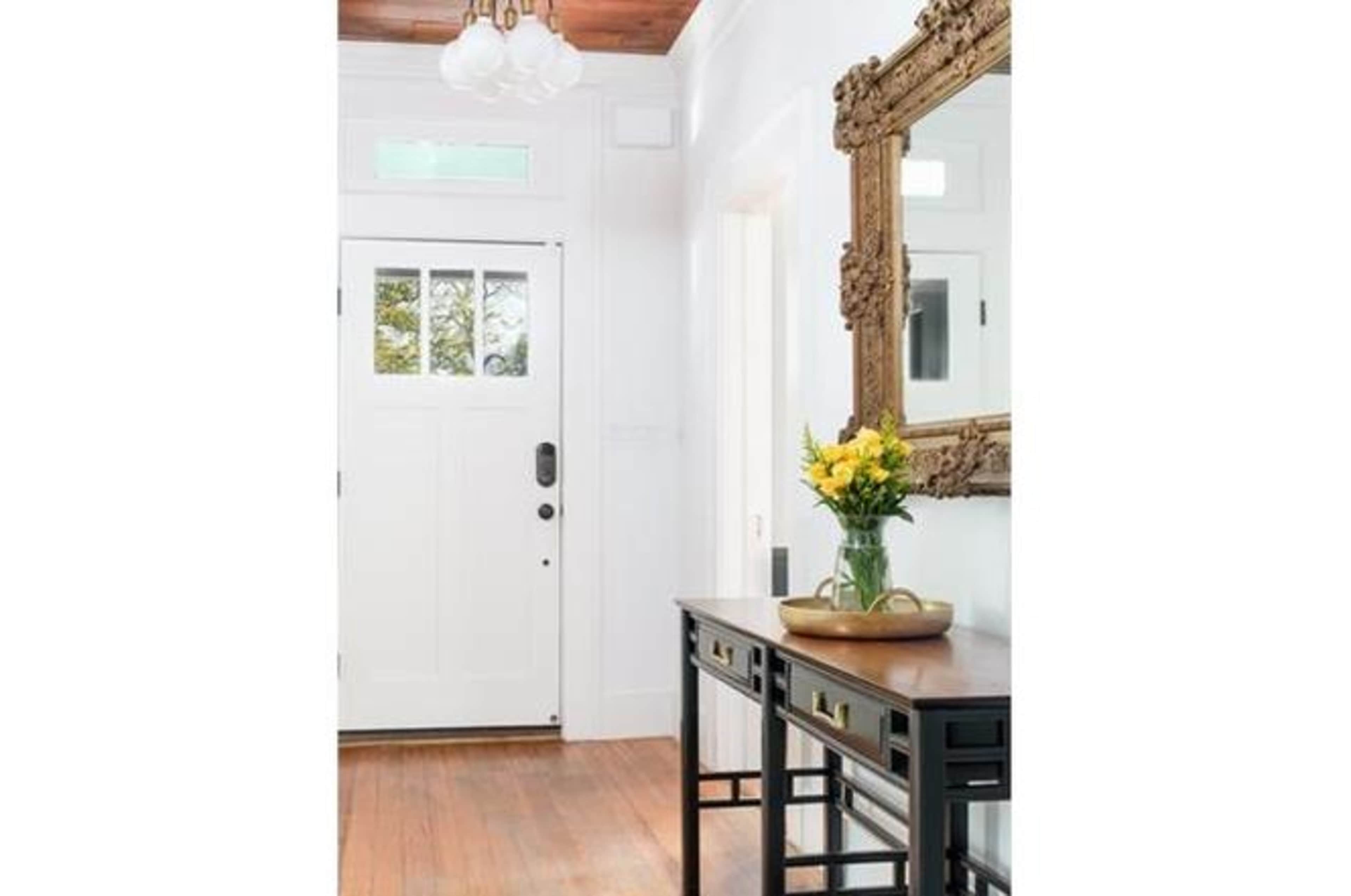 A hallway features a white door, a wooden table with a flower vase, and a decorative mirror on the wall.