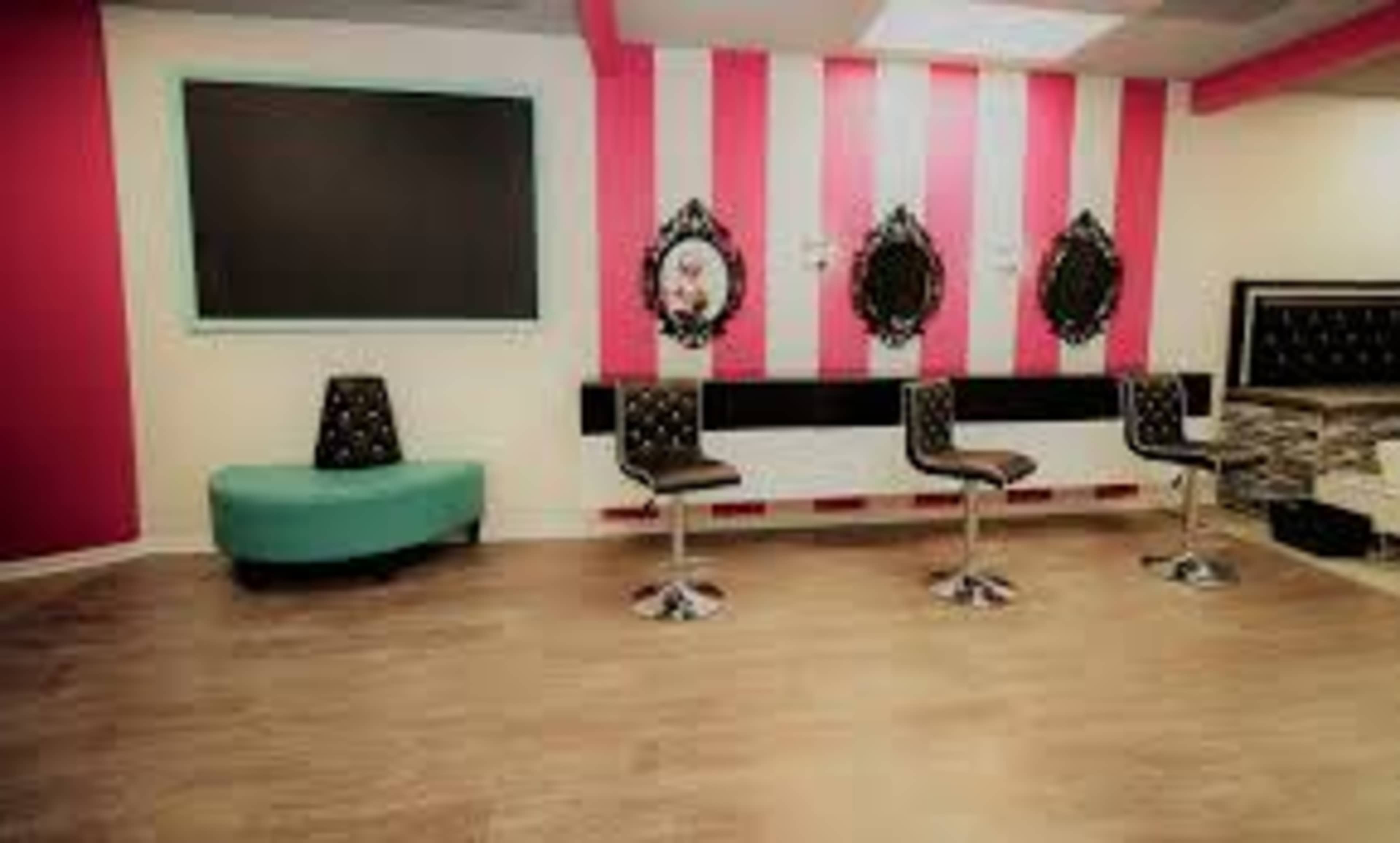 An interior of a salon featuring striped pink walls, a black and white mirror setup, and four bar stools arranged in front of a turquoise seating area.