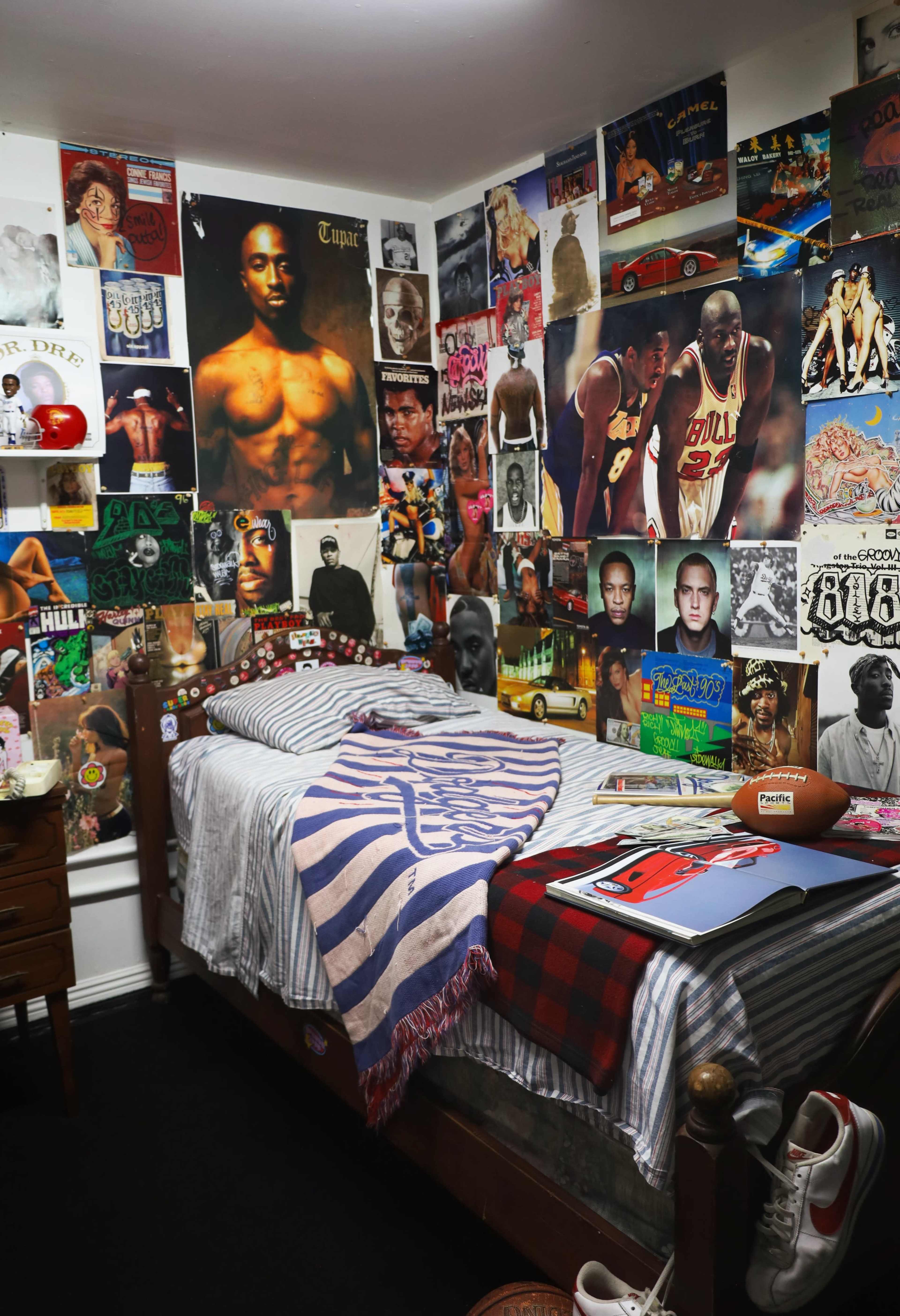 The image shows a bedroom with a bed covered in a striped blanket, surrounded by walls adorned with various posters and memorabilia, including images of musicians and athletes.