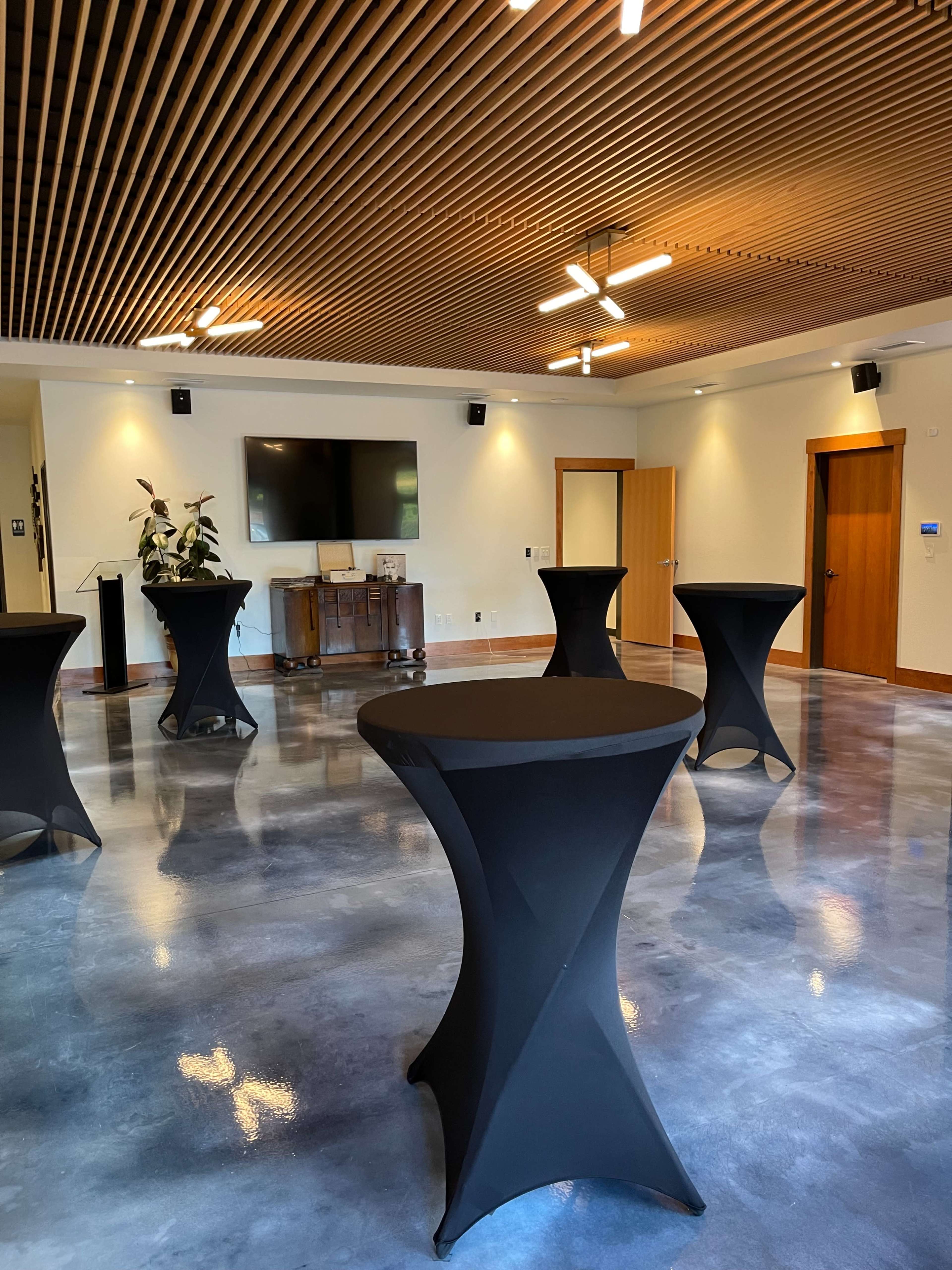 A spacious event room with polished concrete floors, featuring several black cocktail tables arranged in the center and a television mounted on the wall.