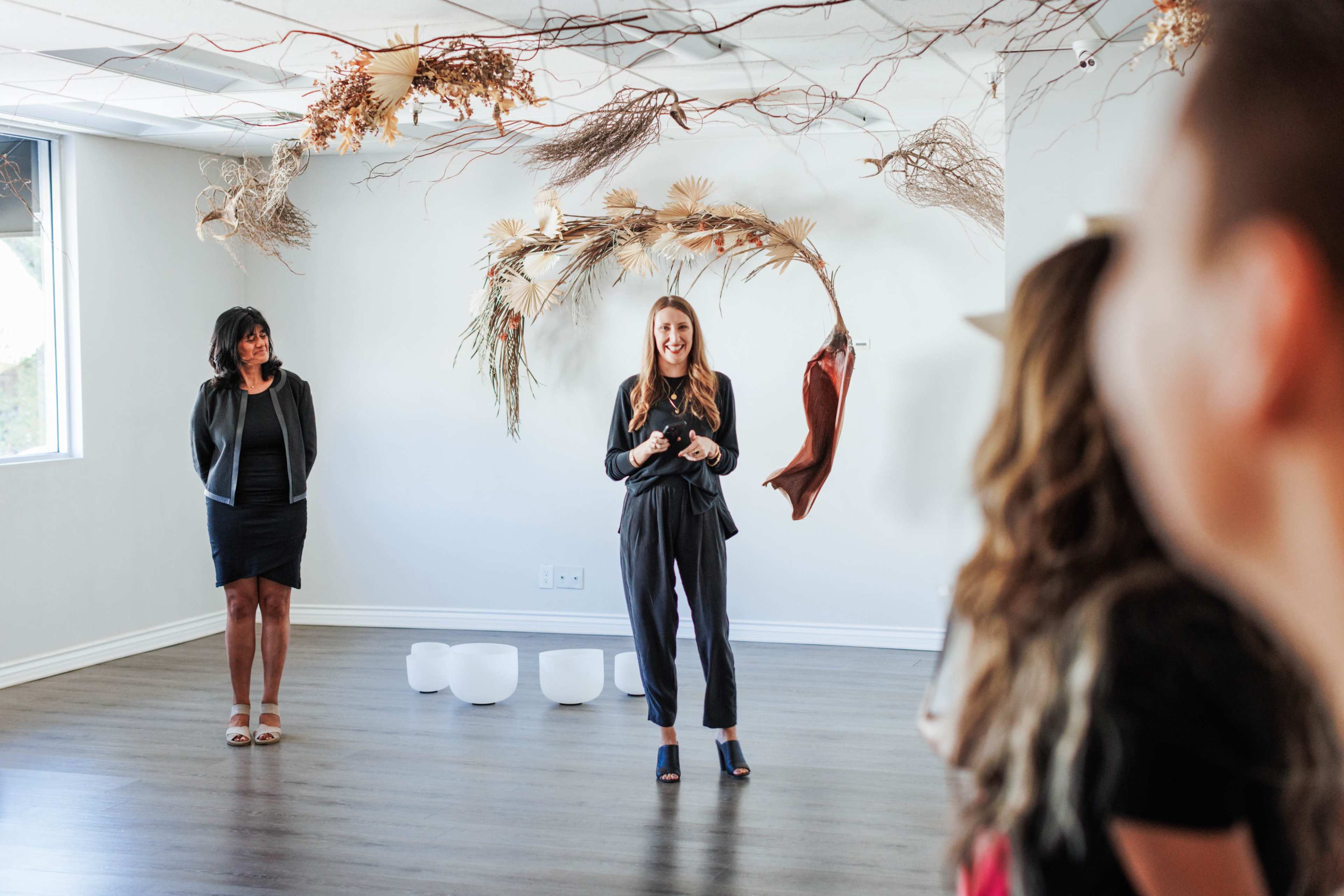 A woman in a black outfit stands speaking in front of a small audience, while another woman stands to her left, with decorative branches and materials hanging from the ceiling.