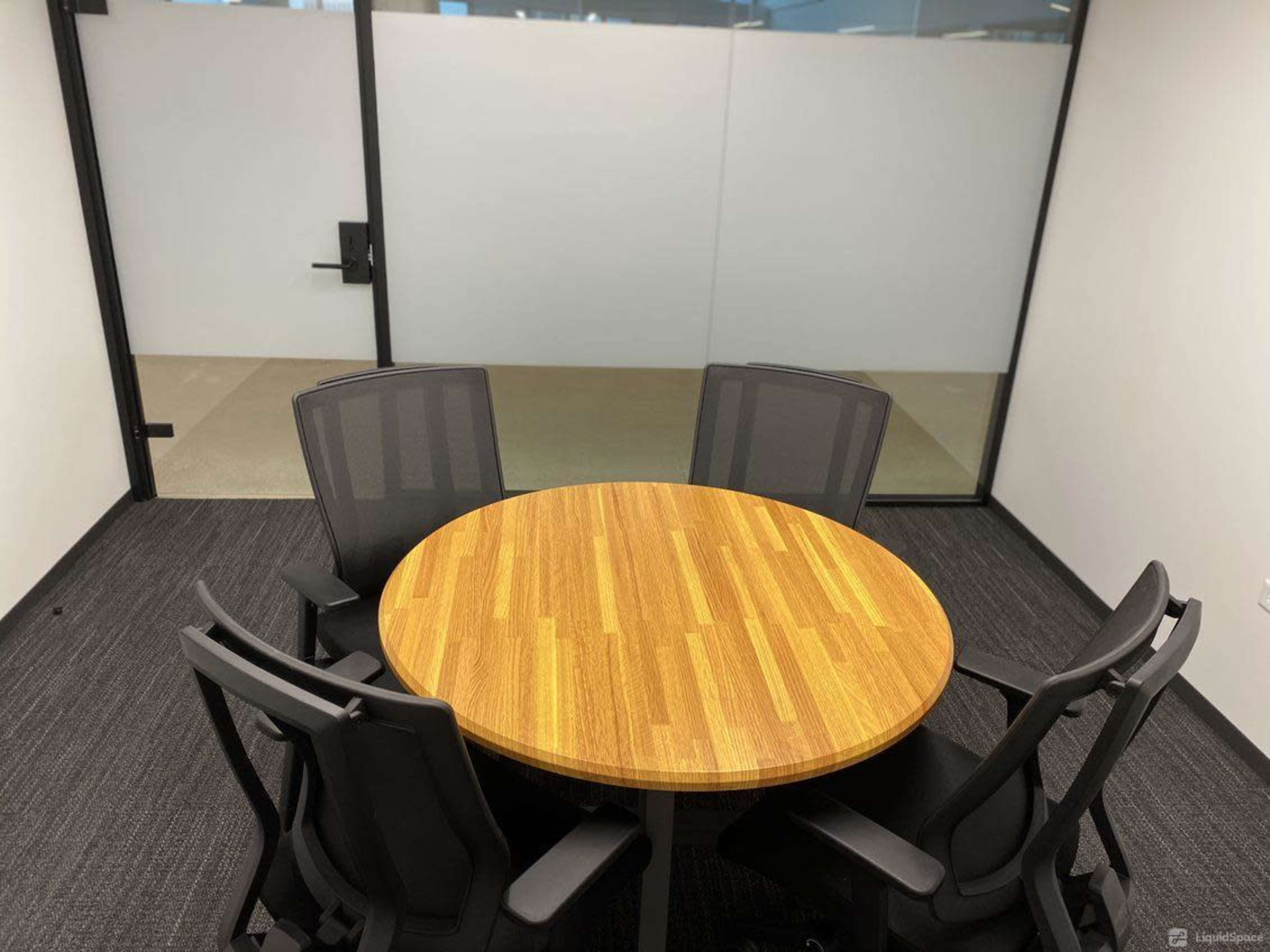 A round wooden table surrounded by four black mesh chairs in a minimalist office space.