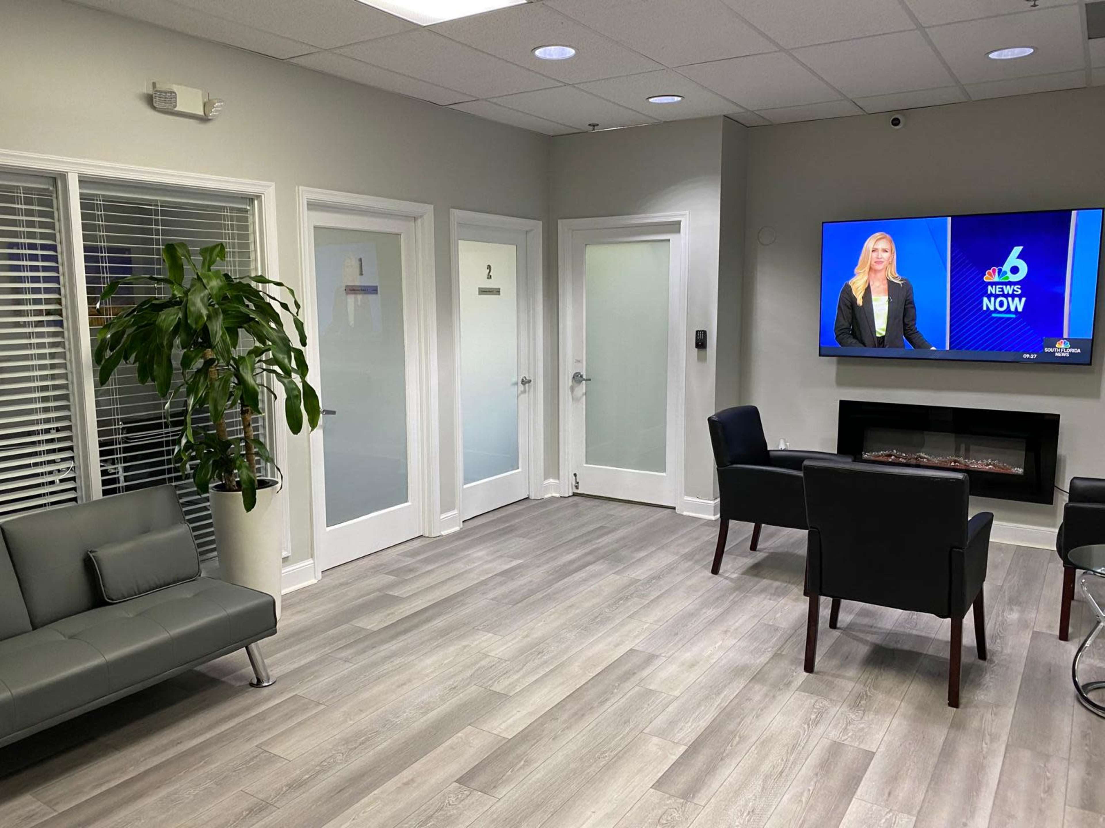The image shows a waiting area with a gray sofa, two black chairs, a large television displaying news, and glass-paneled doors leading to private rooms.