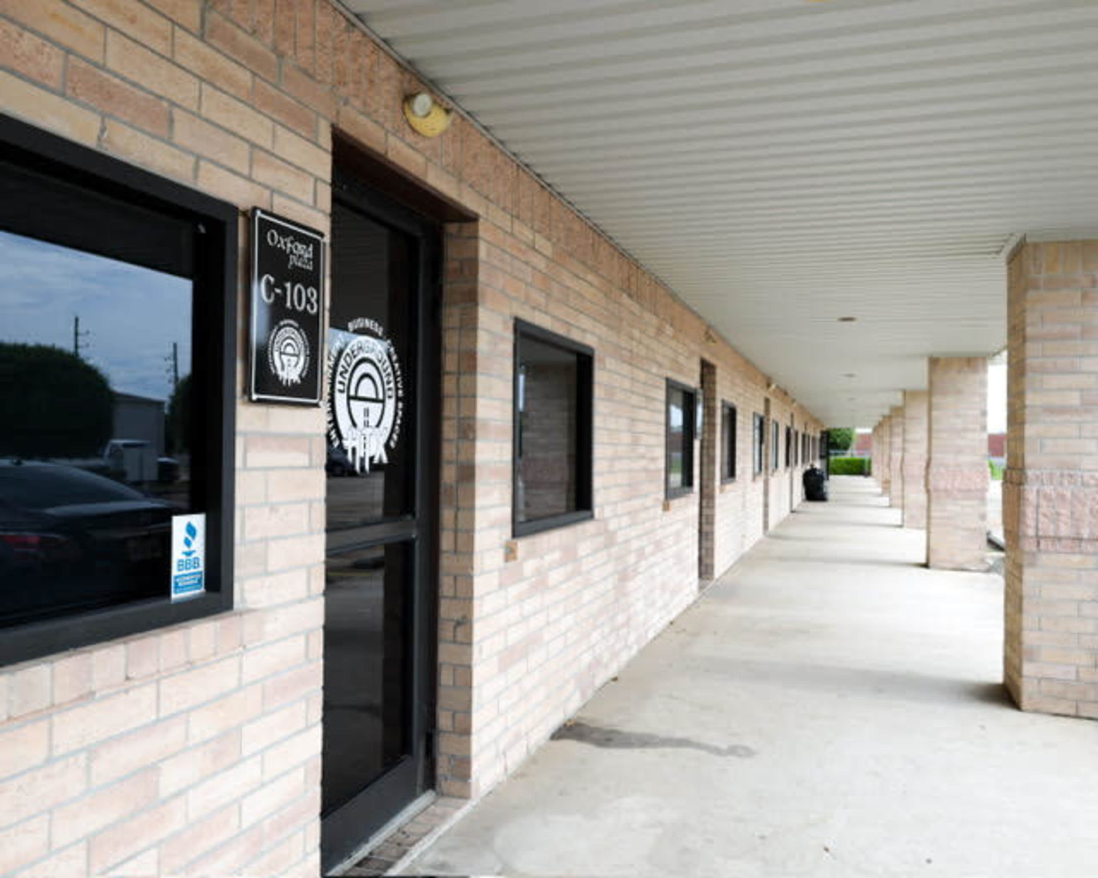 A brick building with a covered walkway and multiple windows, featuring a door labeled "C-103."