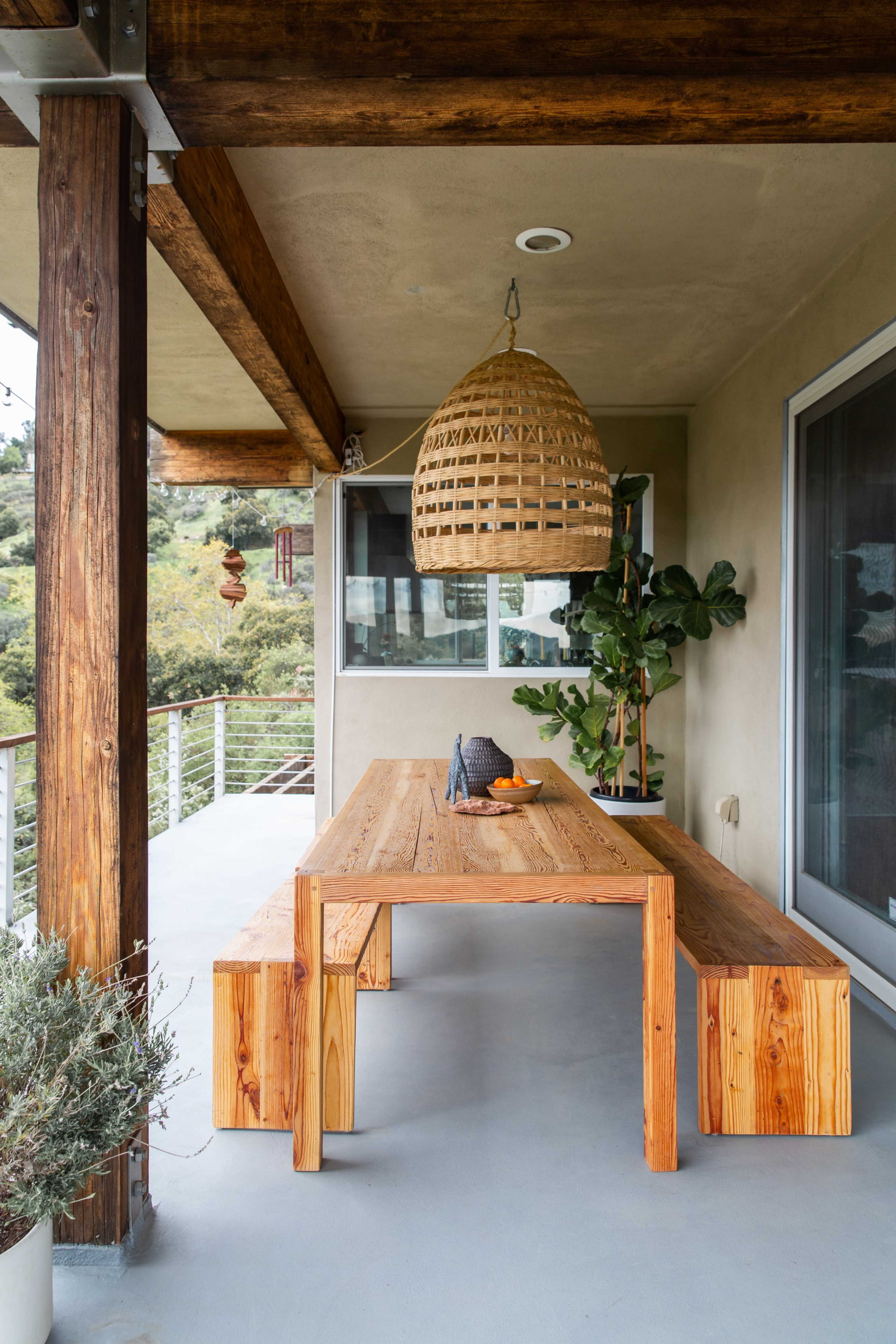 A wooden table with benches sits on a patio under a woven pendant light, surrounded by greenery and a building with large windows.