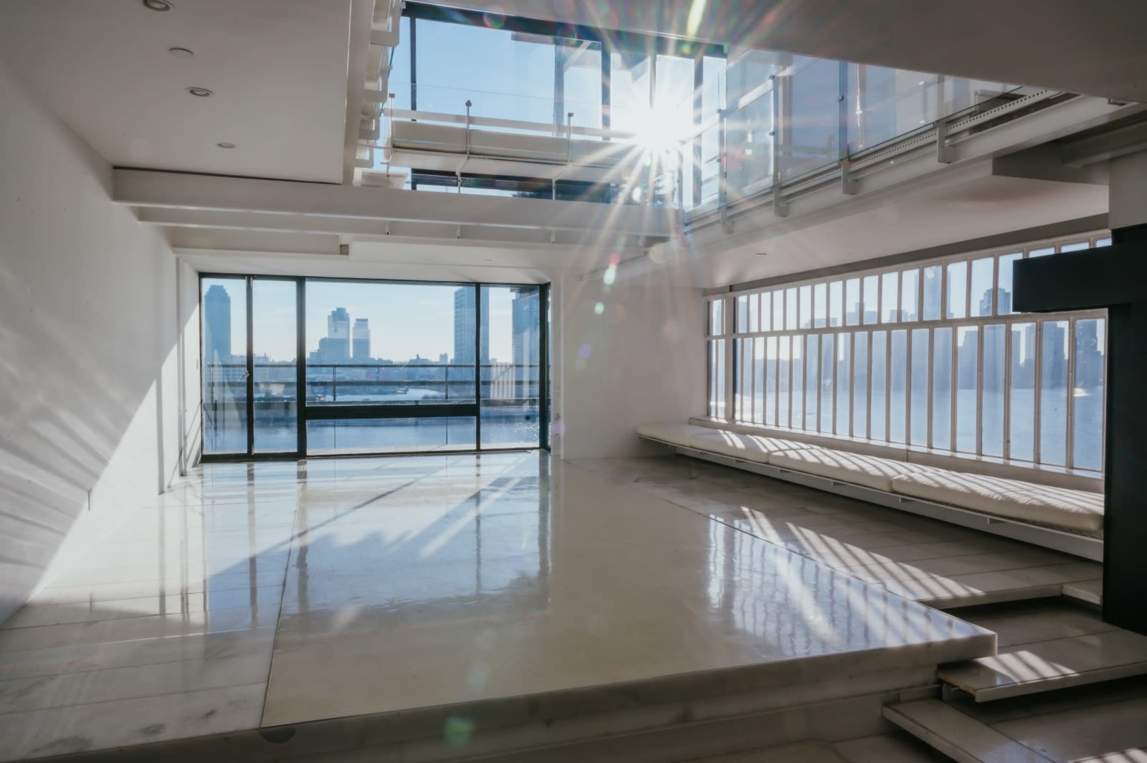 The image depicts a spacious, modern interior with large windows allowing natural light to illuminate the room and a view of a river and skyline beyond.