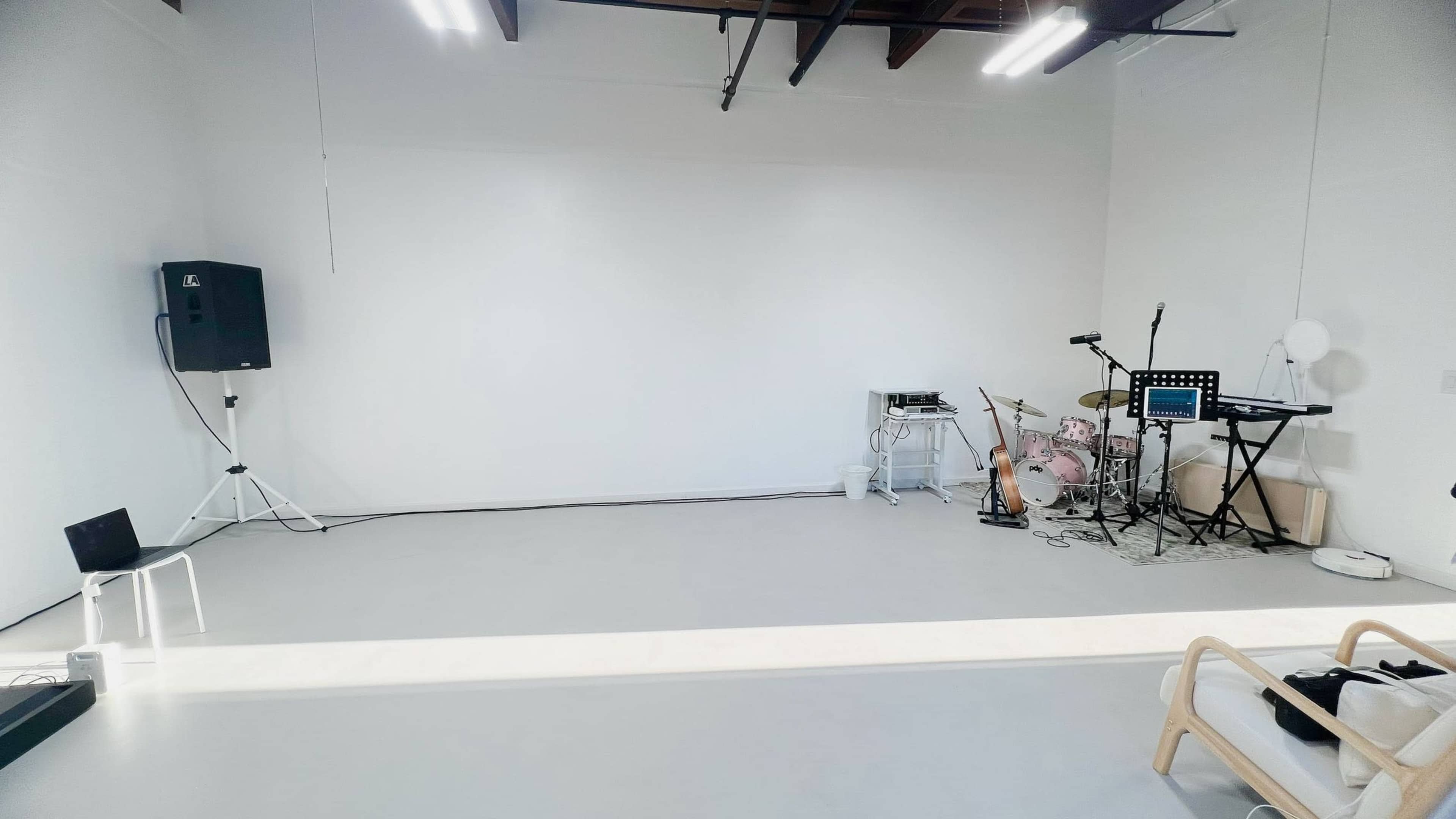 The image shows an empty white room with a small stage setup, including a drum kit, keyboard, and audio equipment.