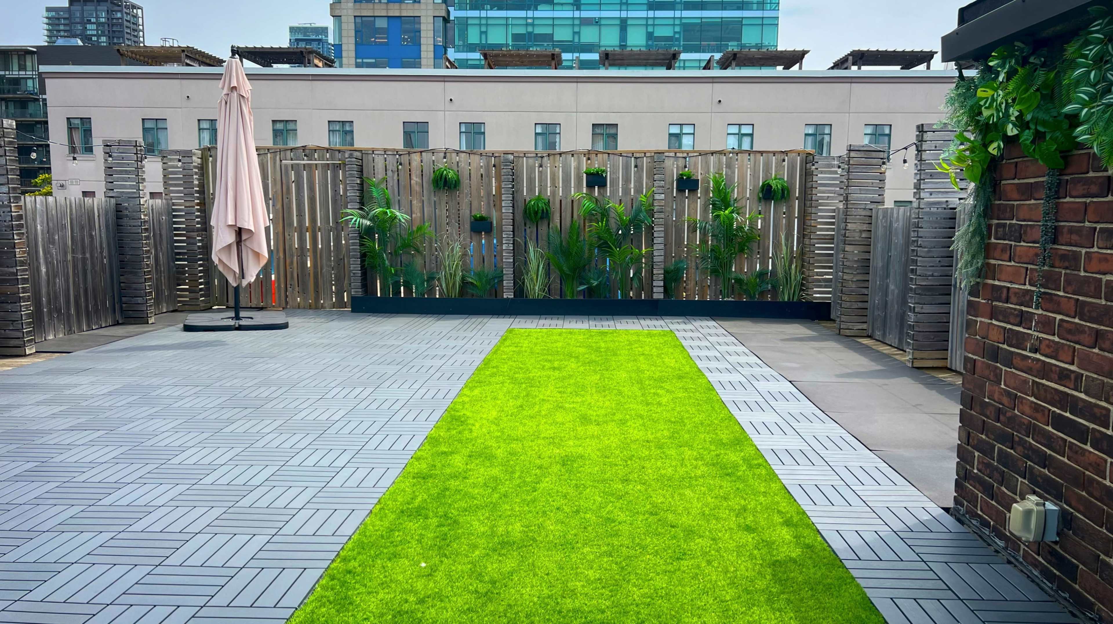 The image shows a rooftop space featuring a green artificial grass pathway bordered by wooden fencing and potted plants, with an umbrella and modern buildings in the background.