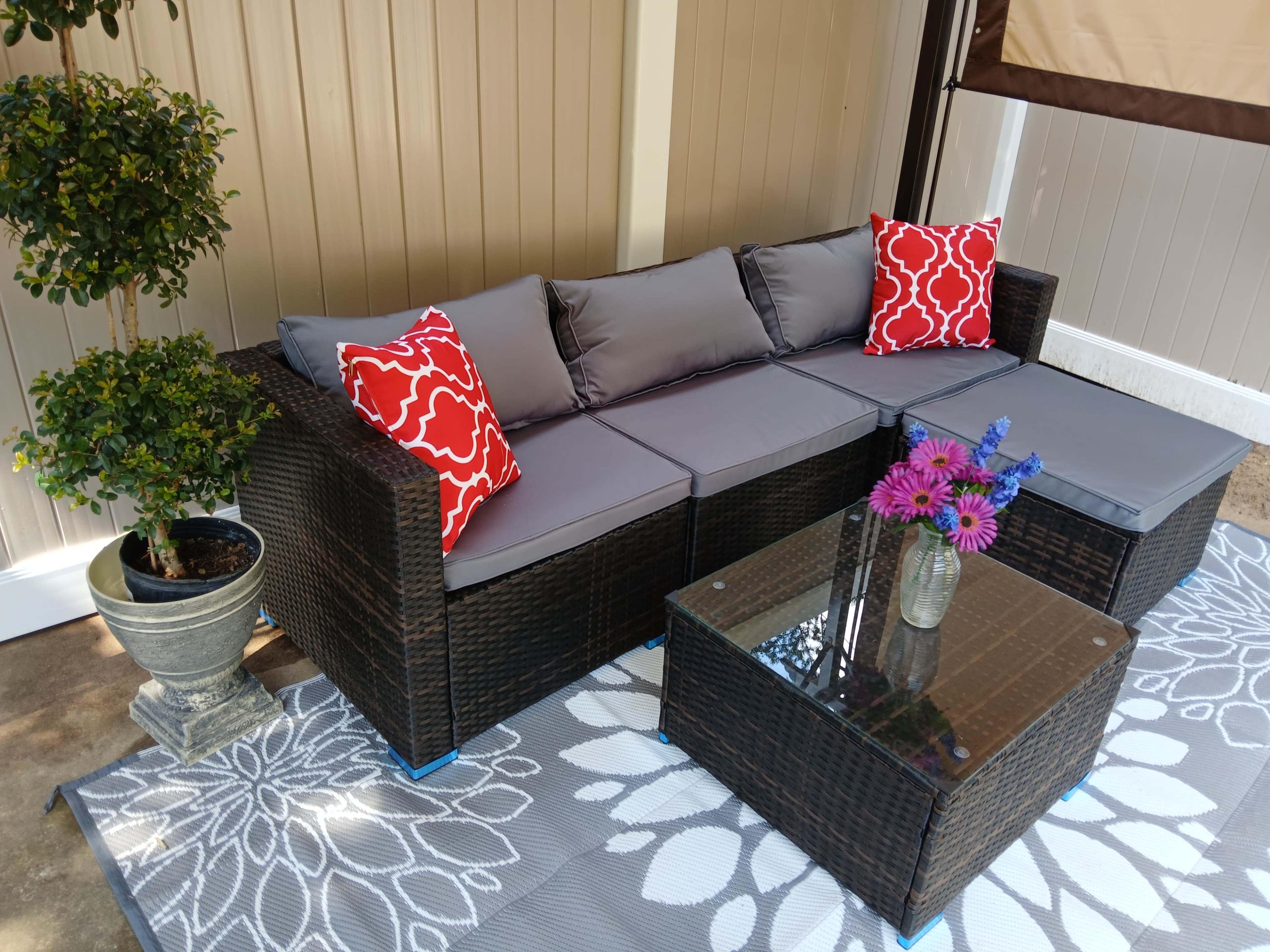 A patio seating area features a dark wicker sectional sofa with gray cushions and red accent pillows, accompanied by a glass-topped coffee table and a potted plant.