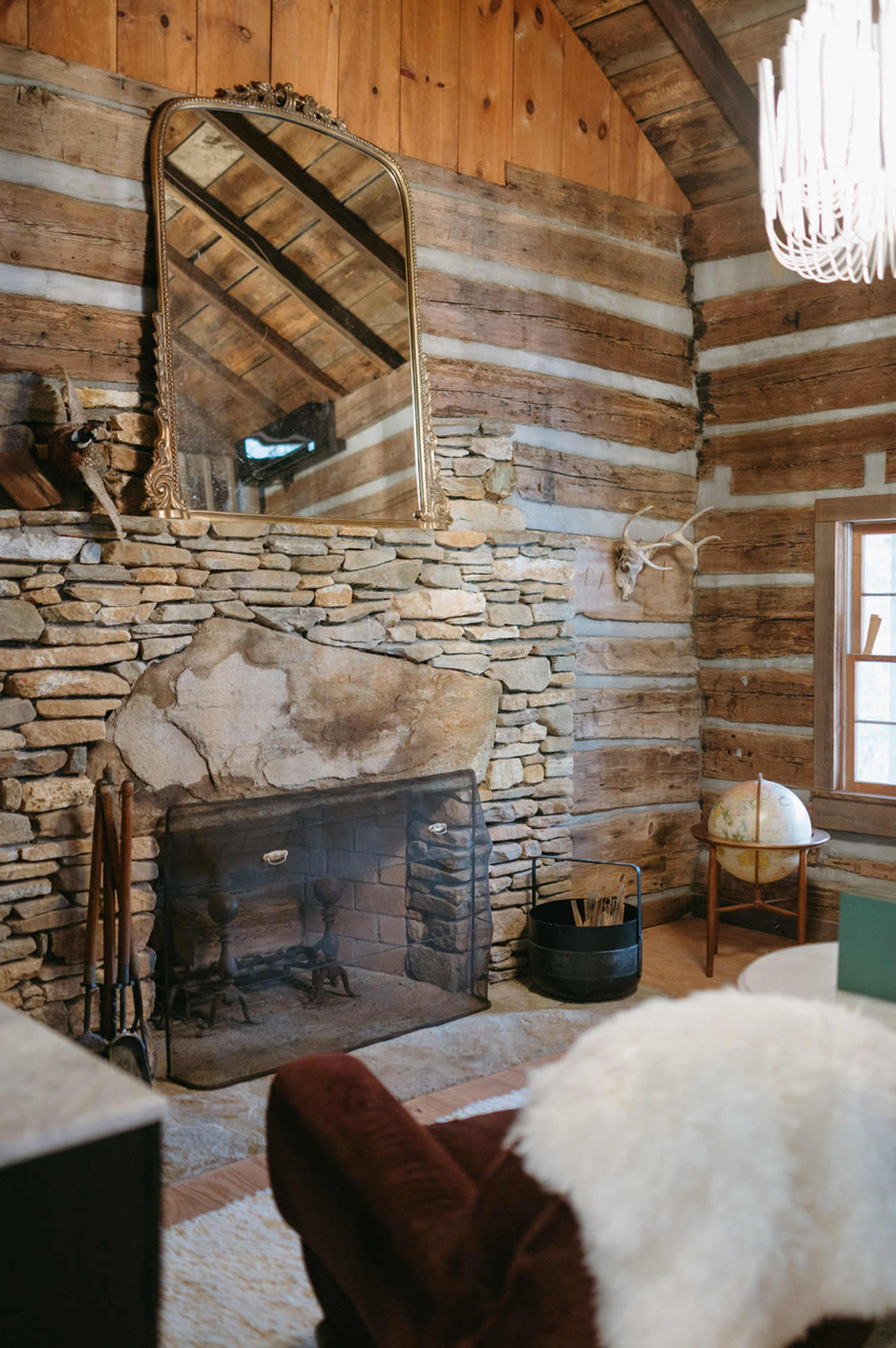 The image shows a rustic living room with a stone fireplace, wooden walls, a large mirror, and a cozy seating area.