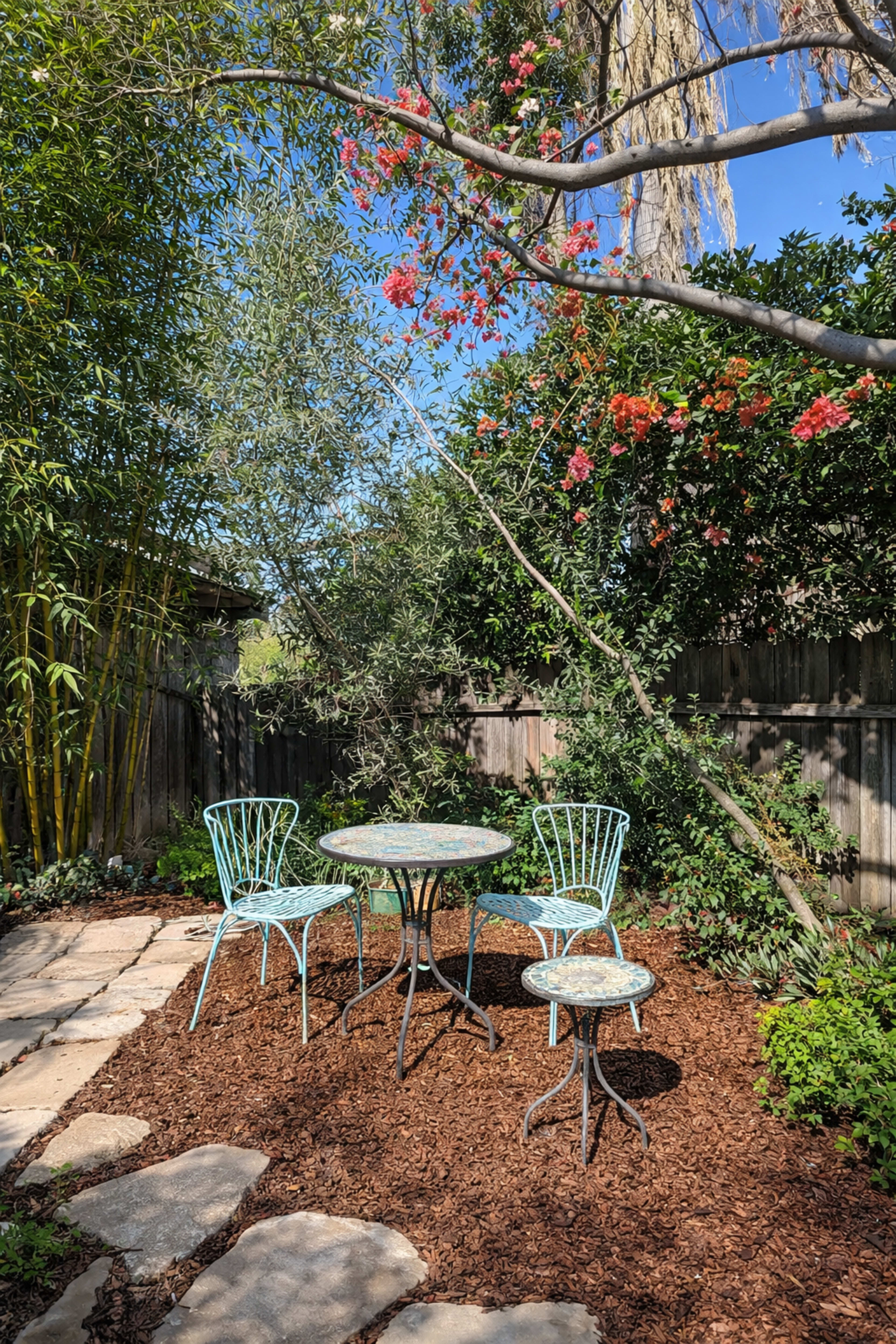 A small, circular outdoor table surrounded by four wire chairs is set on a stone path in a garden with lush greenery and blooming flowers.