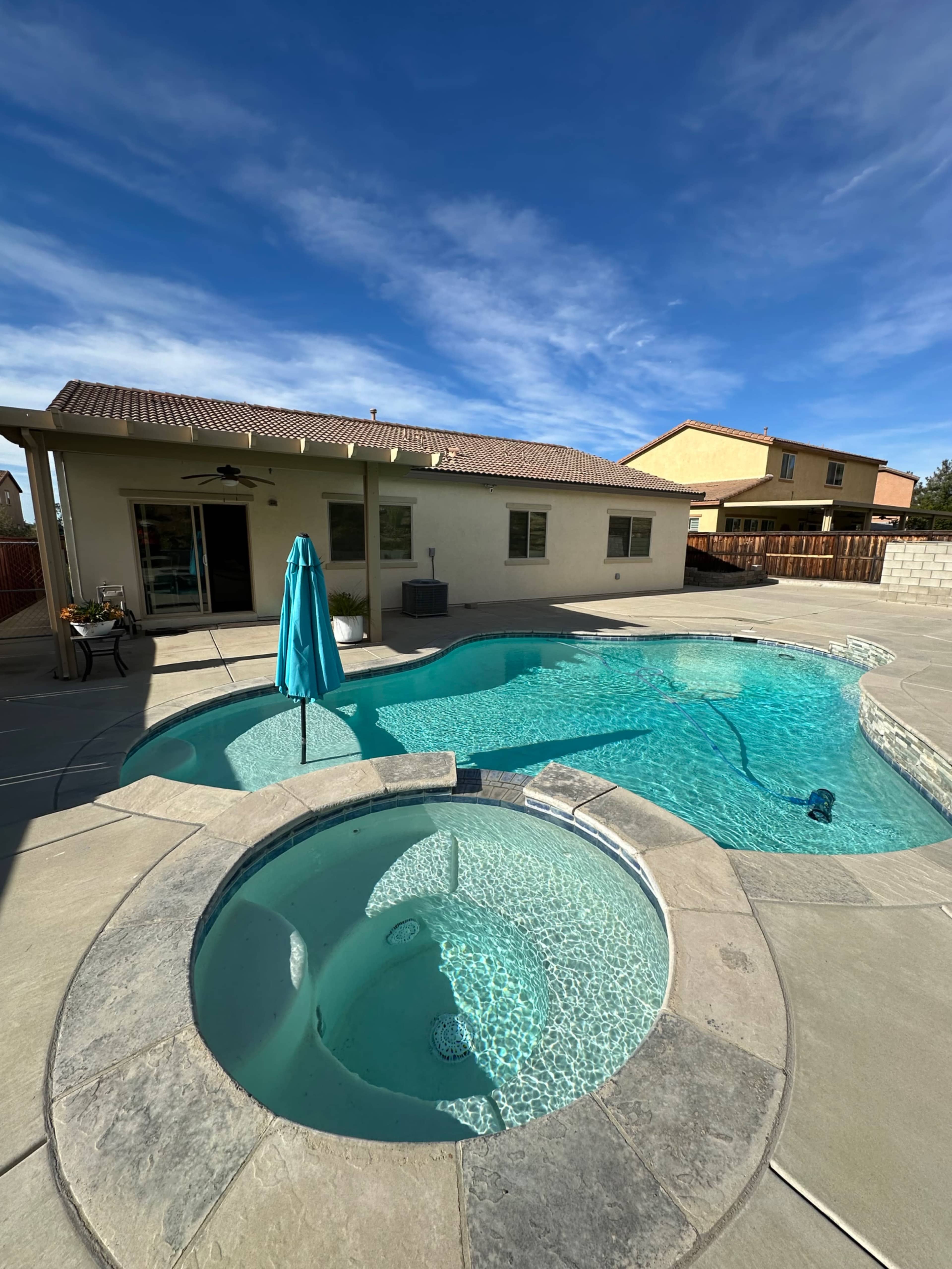The image shows a residential backyard with a swimming pool, a spa, and a house in the background under a clear blue sky.
